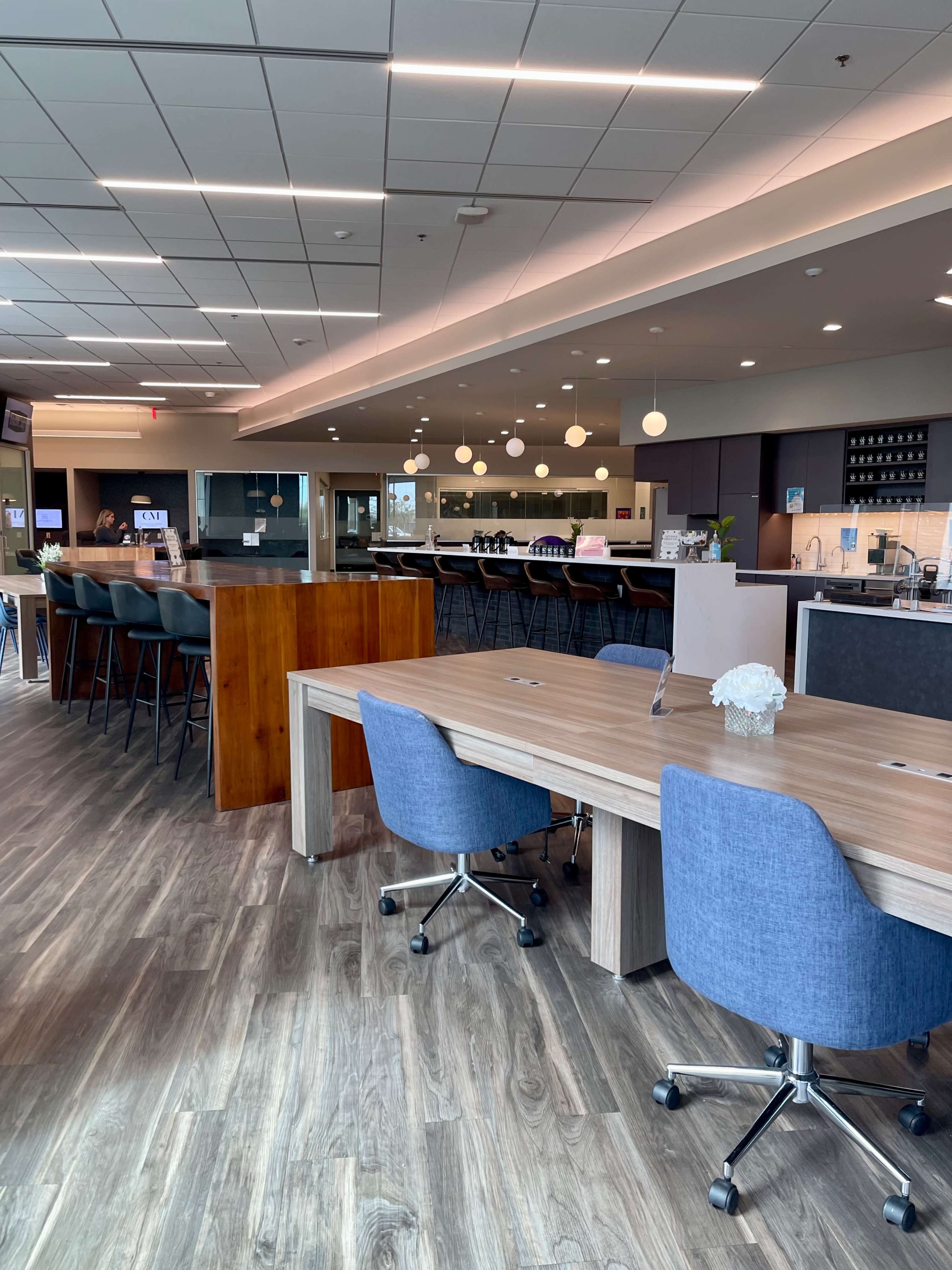 The image shows a modern office space featuring wooden tables with blue swivel chairs, a sleek kitchen area, and pendant lights hanging overhead.