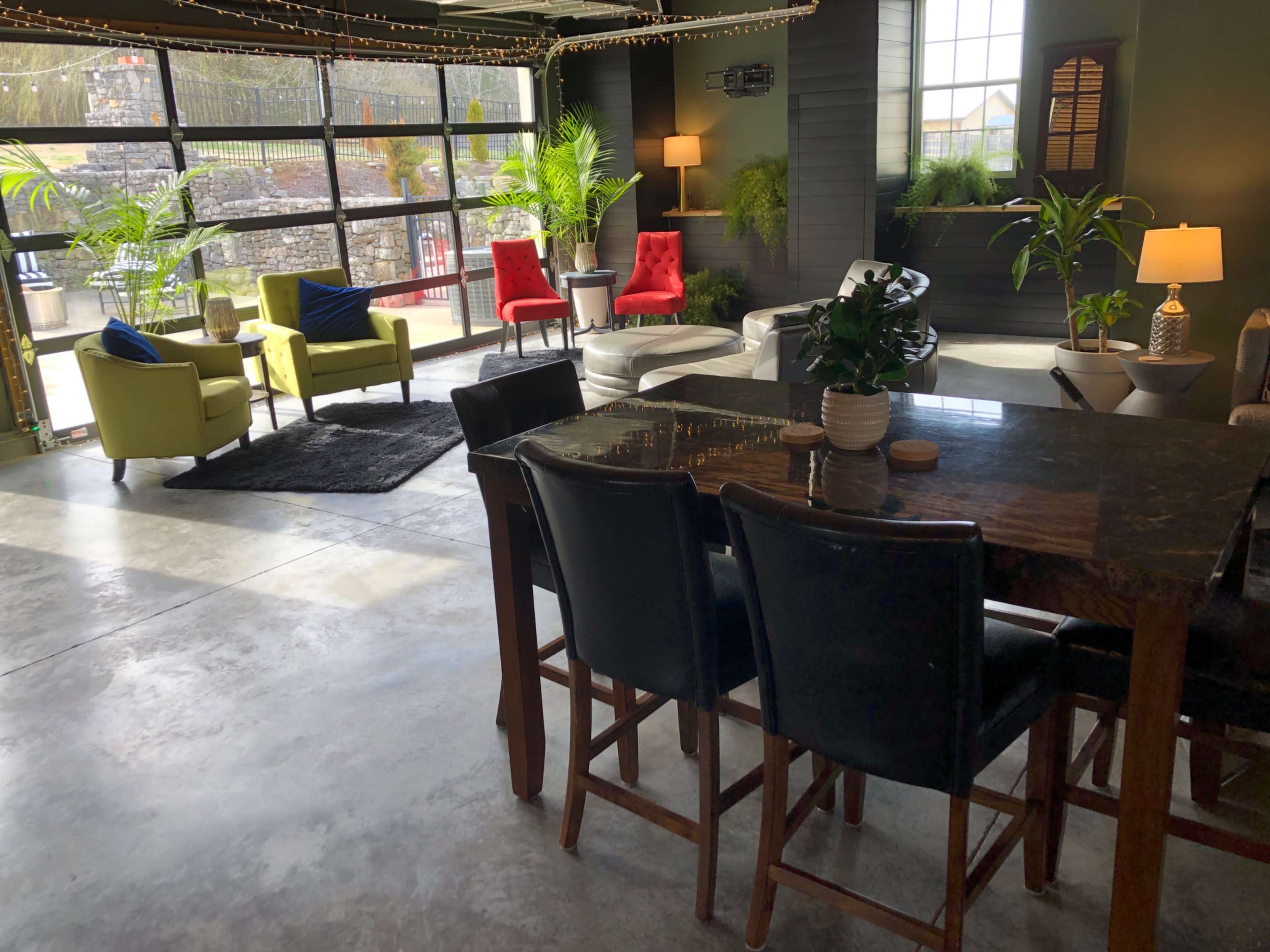 A modern indoor space features a dining area with bar chairs, a table made of dark wood, and a lounge area with various colorful seating arrangements and plants, all illuminated by natural light from large windows.