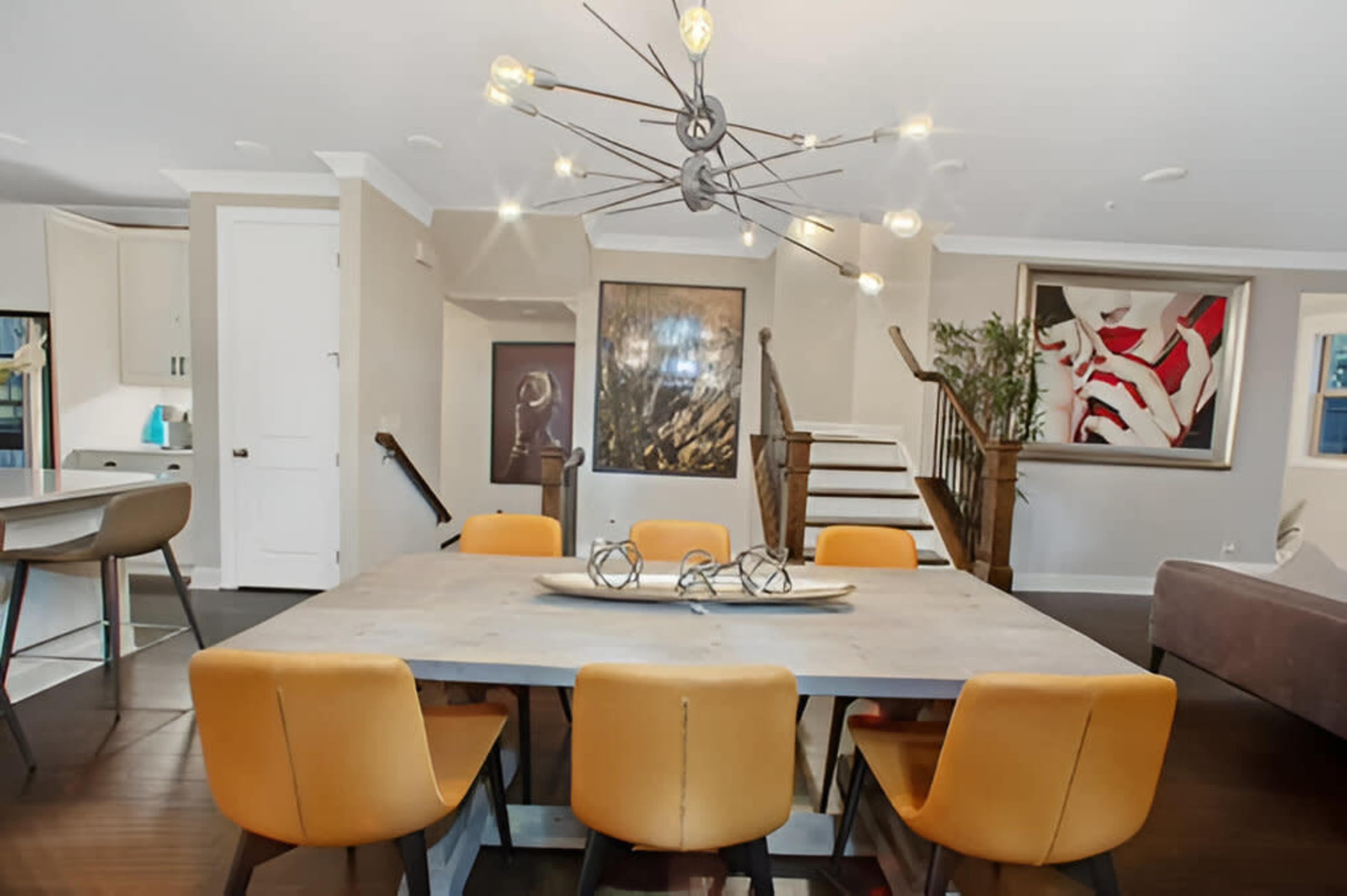 A modern dining area features a large wooden table surrounded by orange chairs, with a striking chandelier above and artwork on the walls.