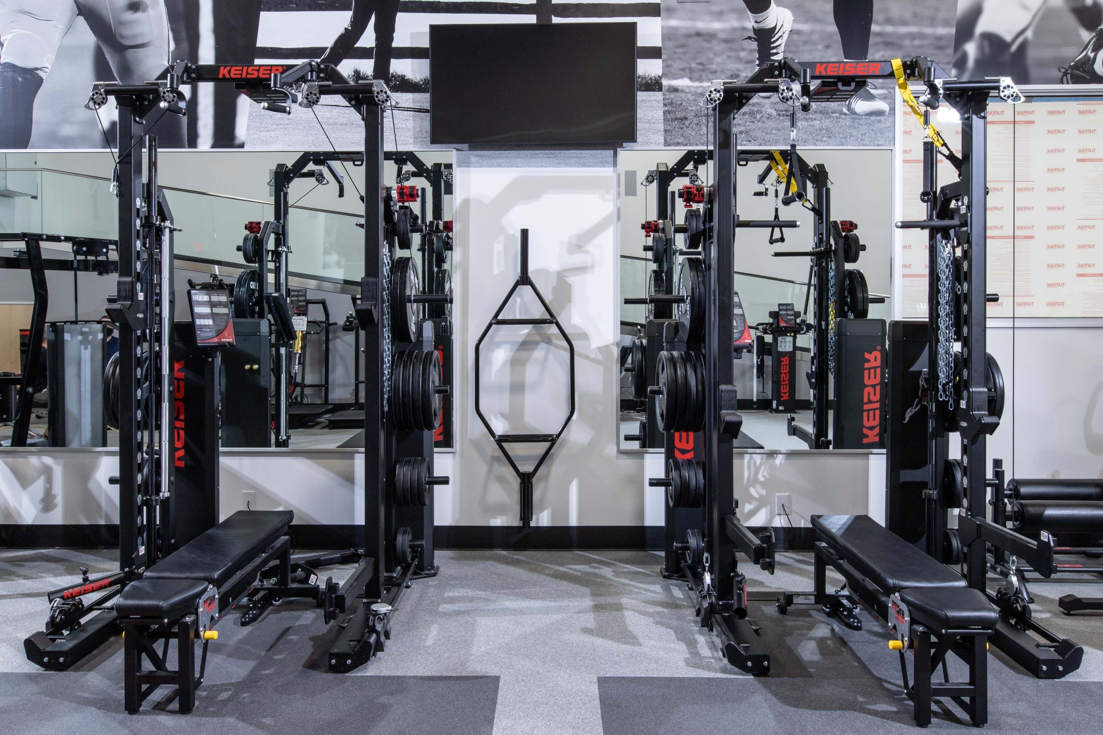 The image shows a modern gym setup featuring two sets of weight training machines positioned back-to-back, with benches and weight plates organized on either side.