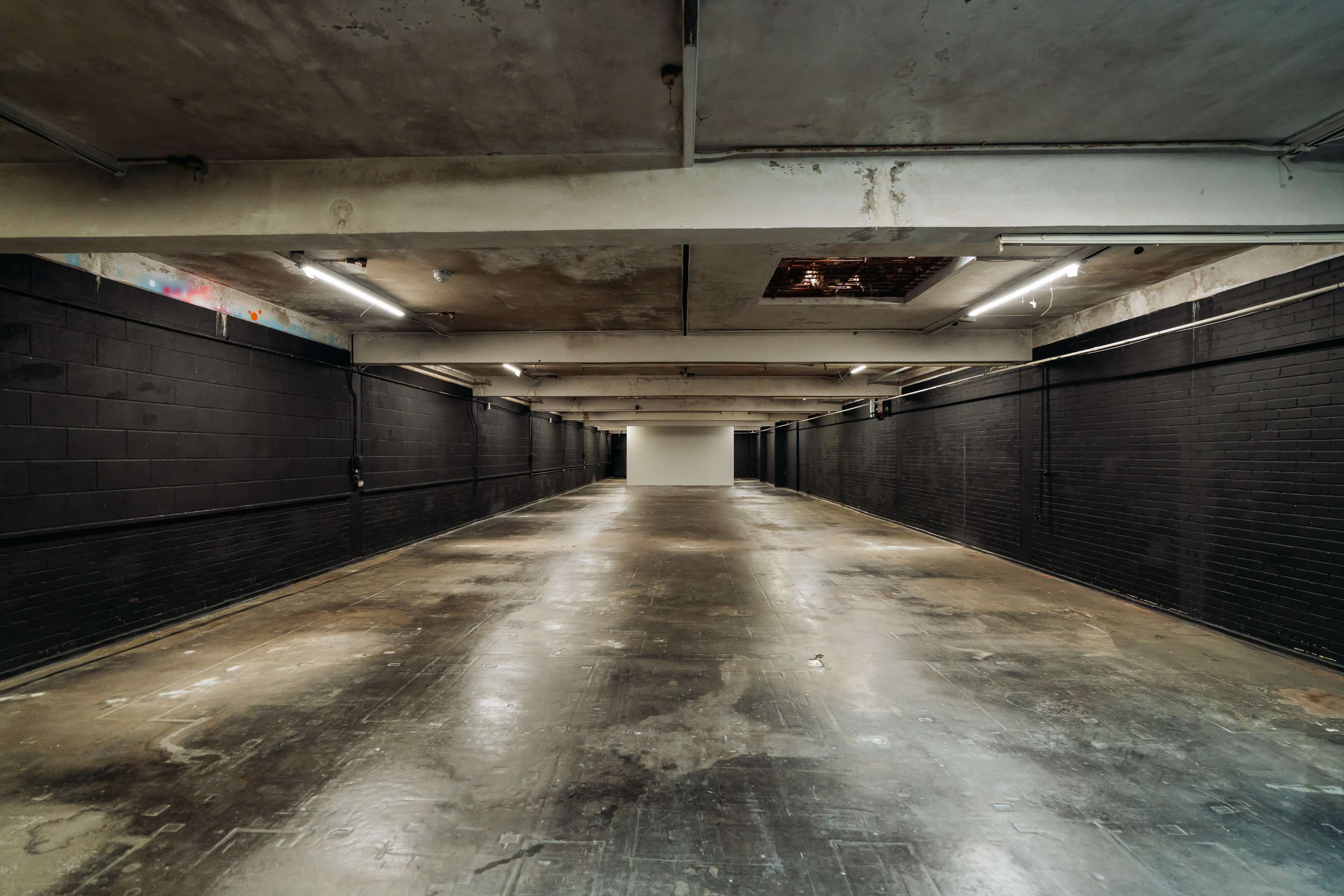 The image shows a long, empty corridor with dark brick walls and a concrete ceiling, illuminated by fluorescent lights.
