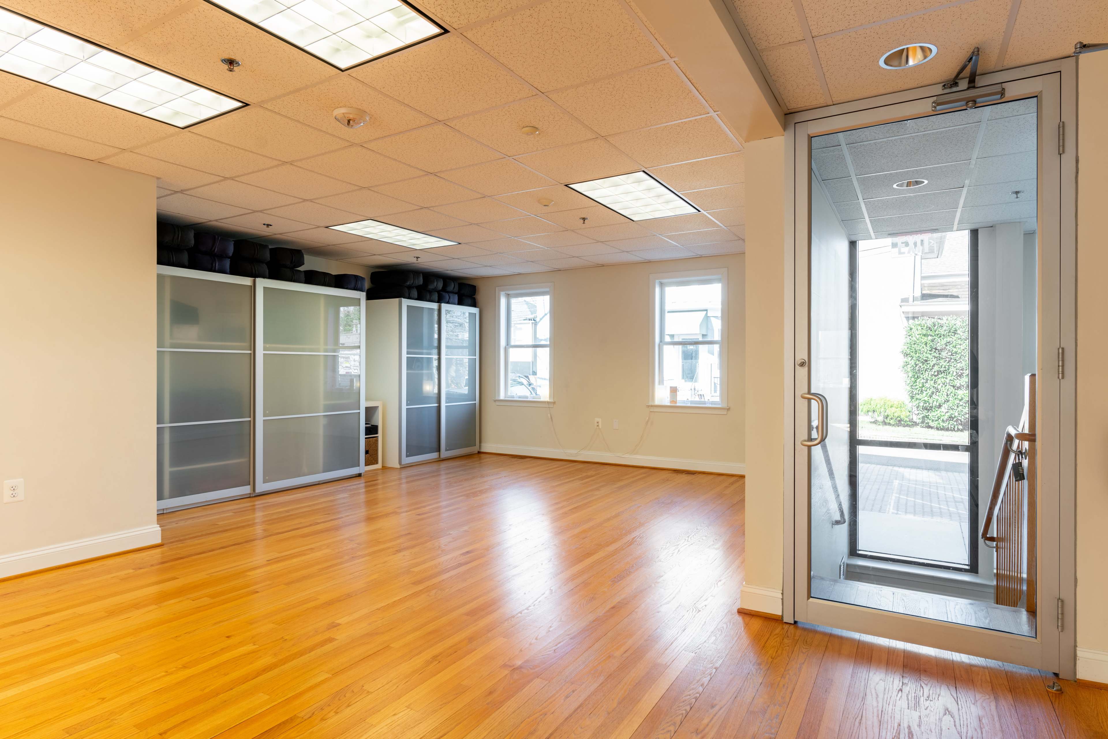 An empty room with hardwood flooring, large windows, and a glass door leading to an exterior walkway.
