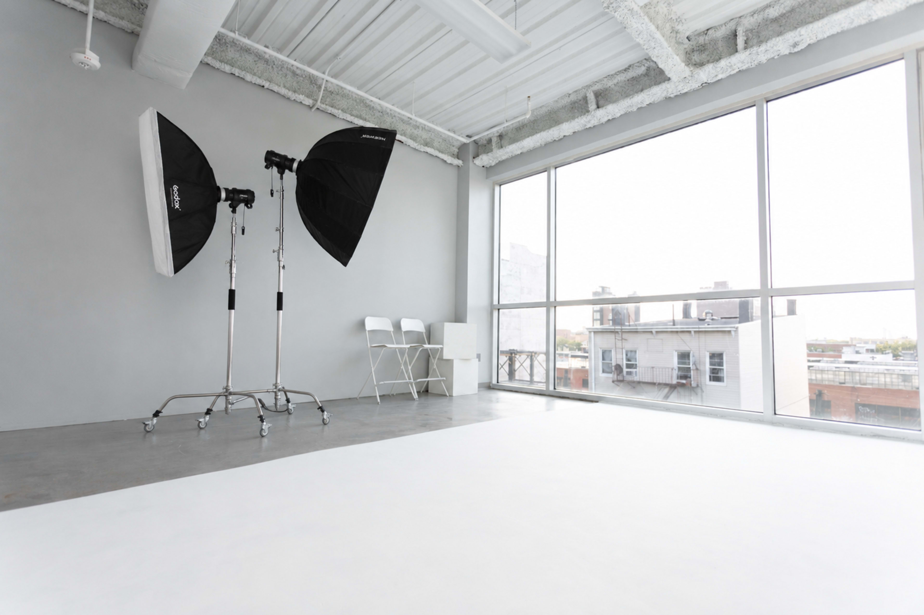 The image shows a spacious photography studio with two large softboxes, a backdrop, and a few chairs near a window overlooking an urban area.
