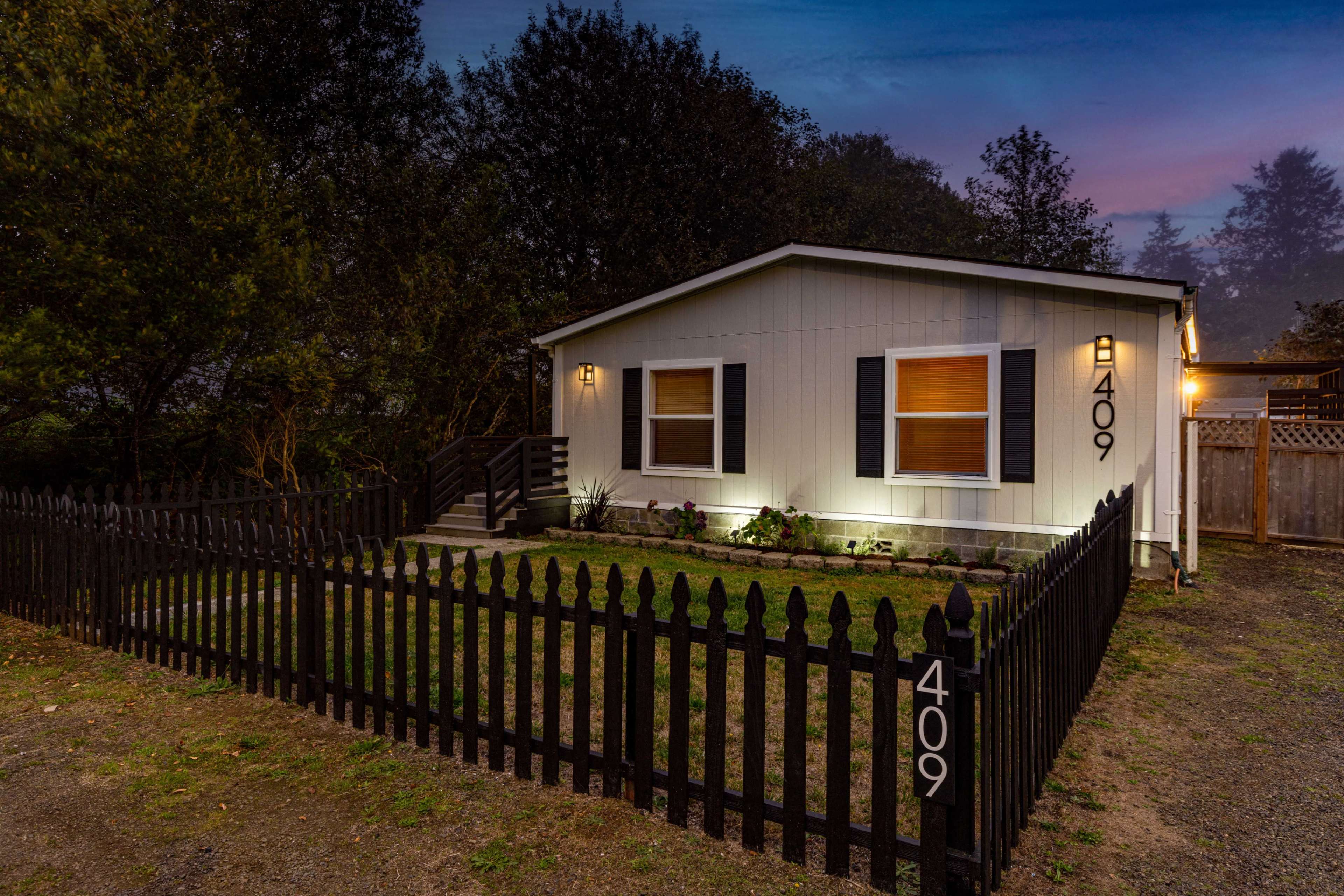 The image shows a small, single-story house with white siding, two windows illuminated at dusk, and a black picket fence enclosing the front yard.
