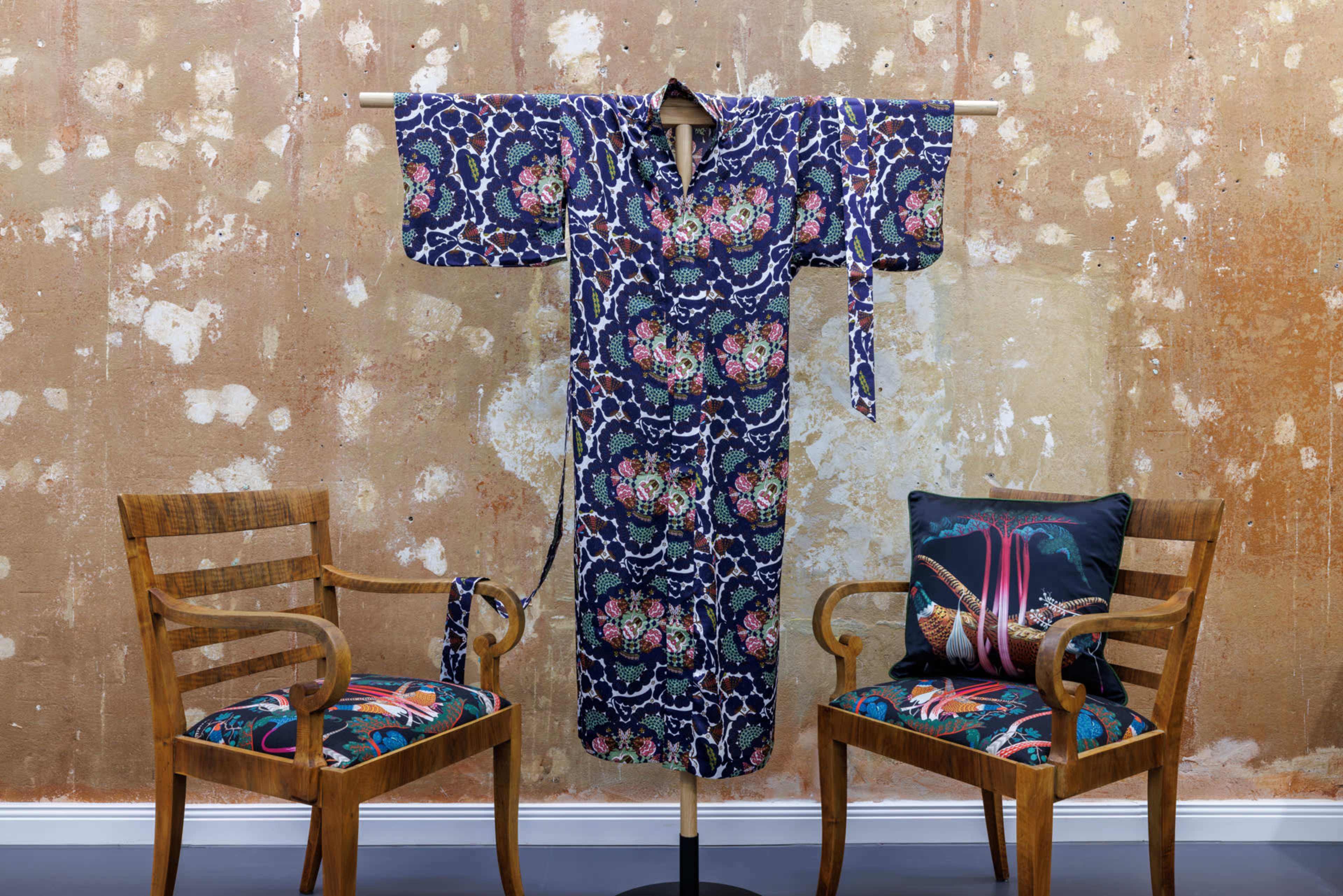 A floral-patterned kimono is displayed on a wooden stand between two wooden chairs featuring colorful seat cushions.