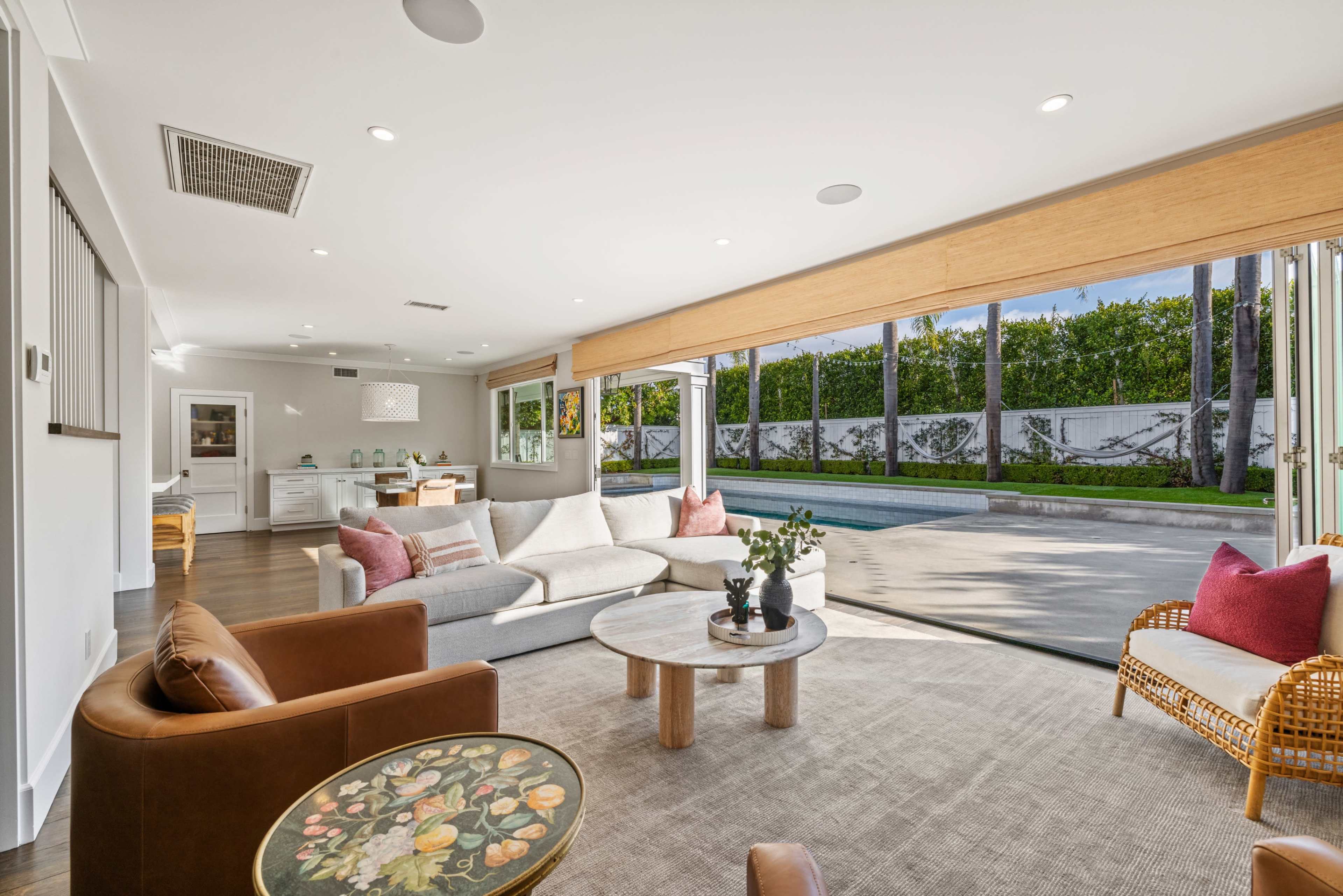 The image shows a modern living room with large windows opening to a backyard pool area, featuring a light-colored sectional sofa, a round coffee table, and various decorative elements.