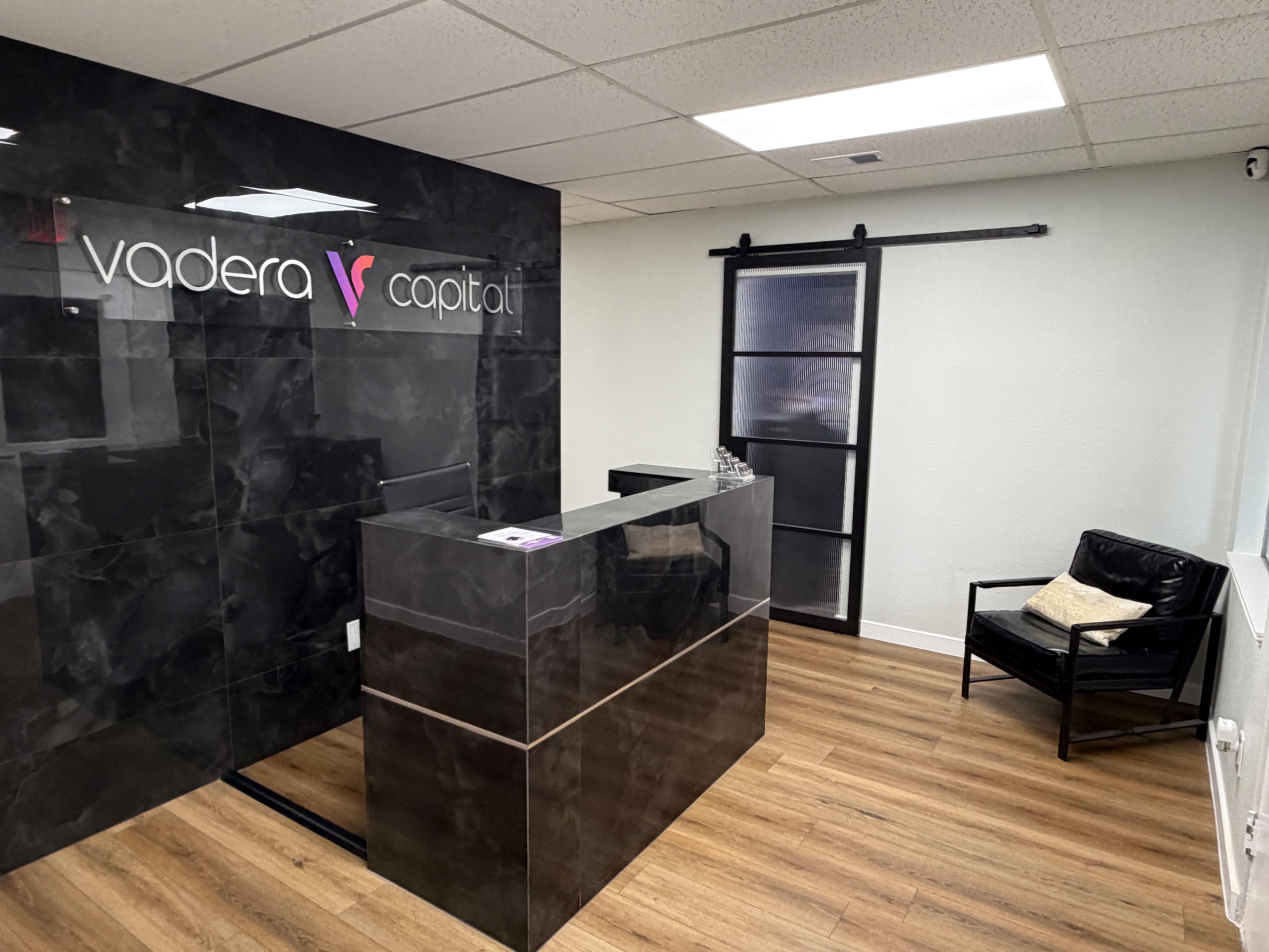 The image shows a modern office reception area featuring a sleek black reception desk with a sign that reads "vadera capital," along with a black chair and a sliding door.