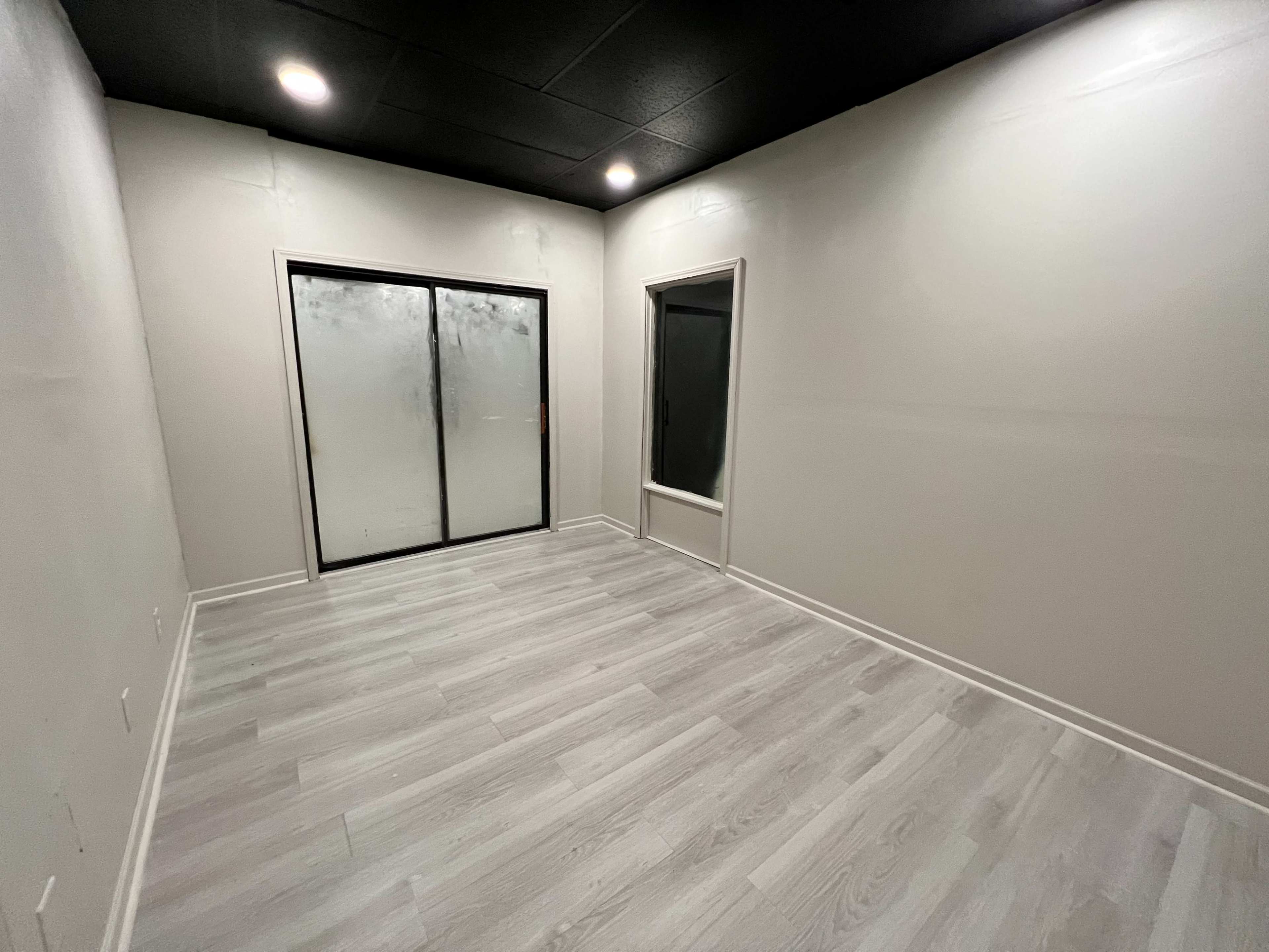 The image shows an empty room with light gray walls, a wood-like floor, and large glass doors.