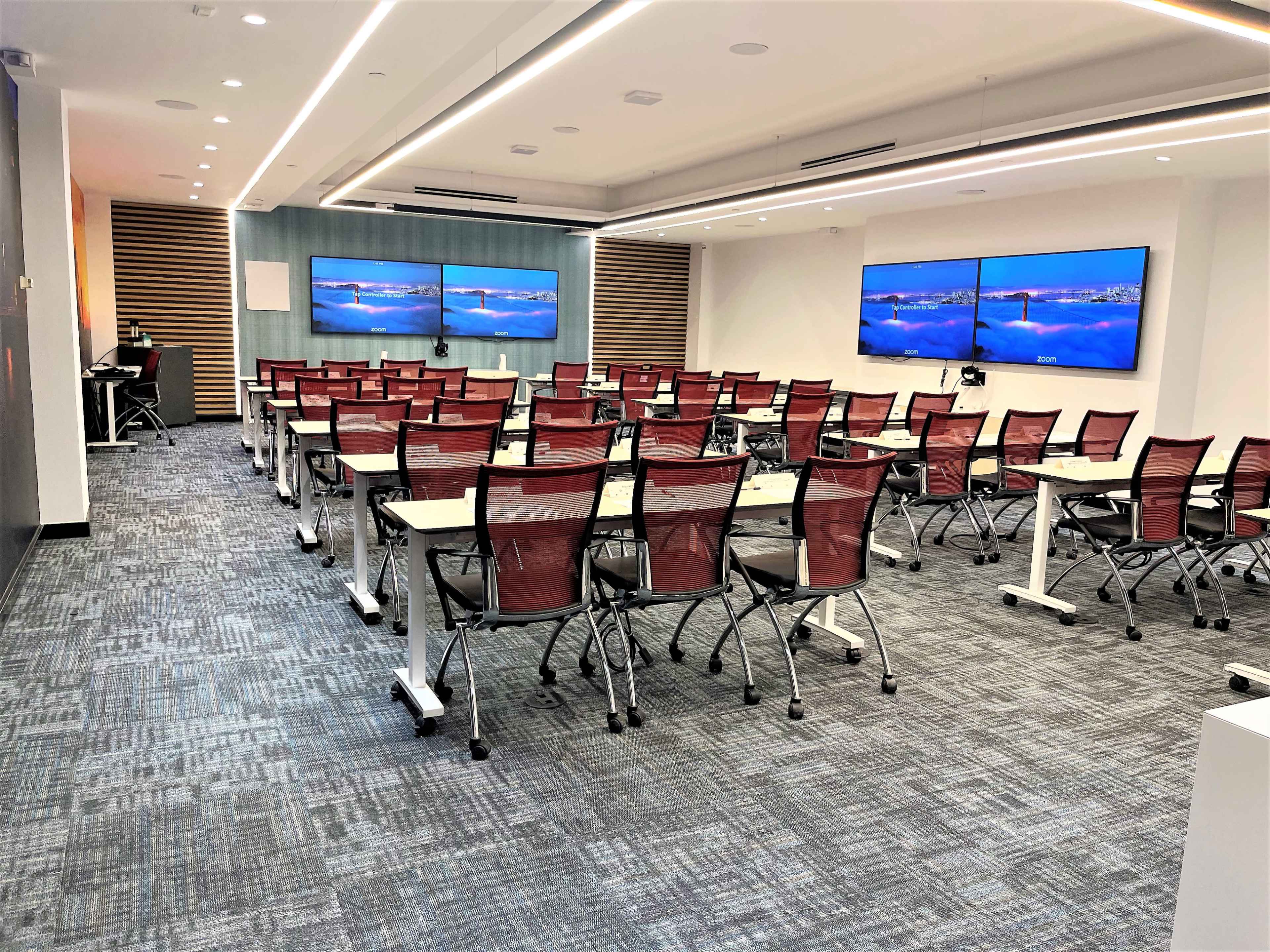 The image shows a modern classroom equipped with rows of desks and chairs facing two large screens on the front wall.