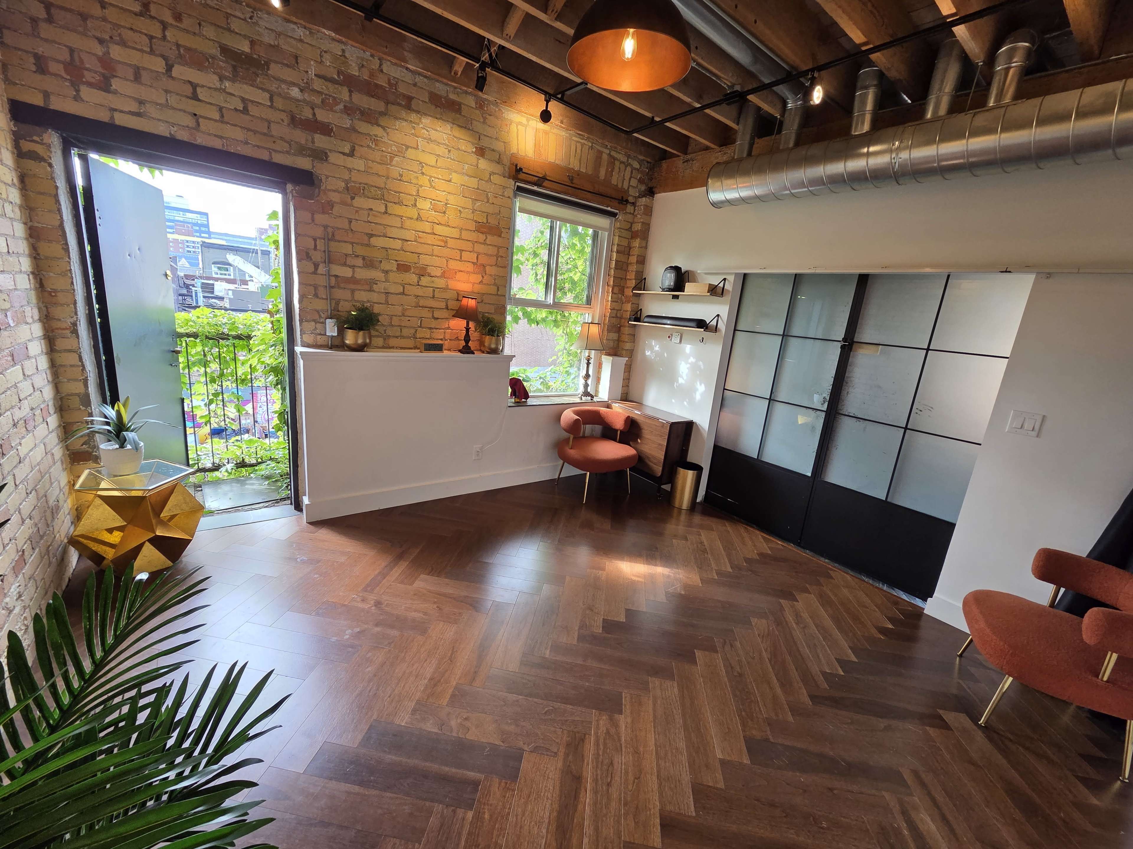 A modern interior space features wooden flooring, a large window with greenery outside, and a combination of sleek furniture, including two chairs and a countertop area.