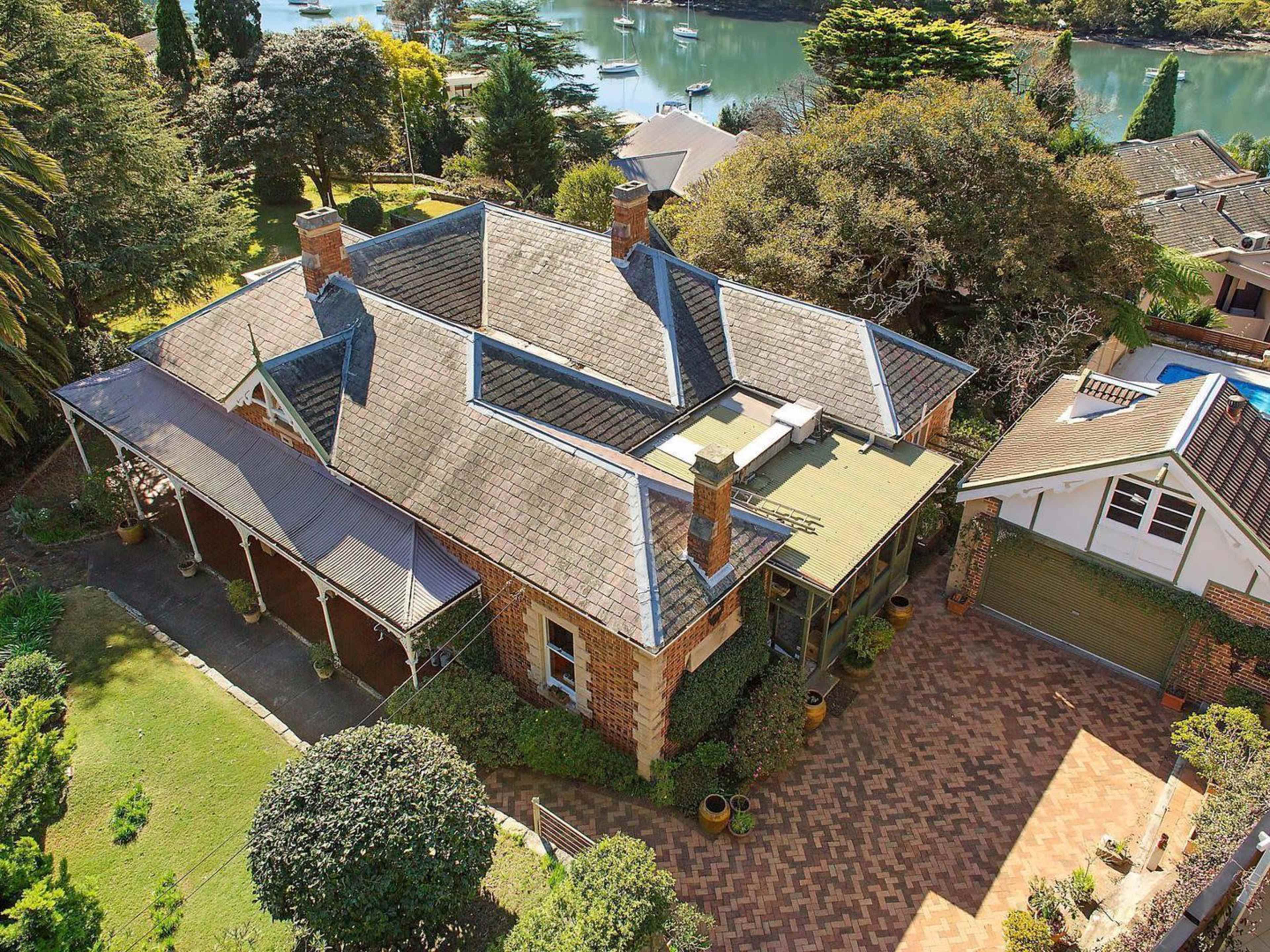 The image shows a large, two-story brick house with multiple gables, surrounded by greenery and a view of a body of water in the background.