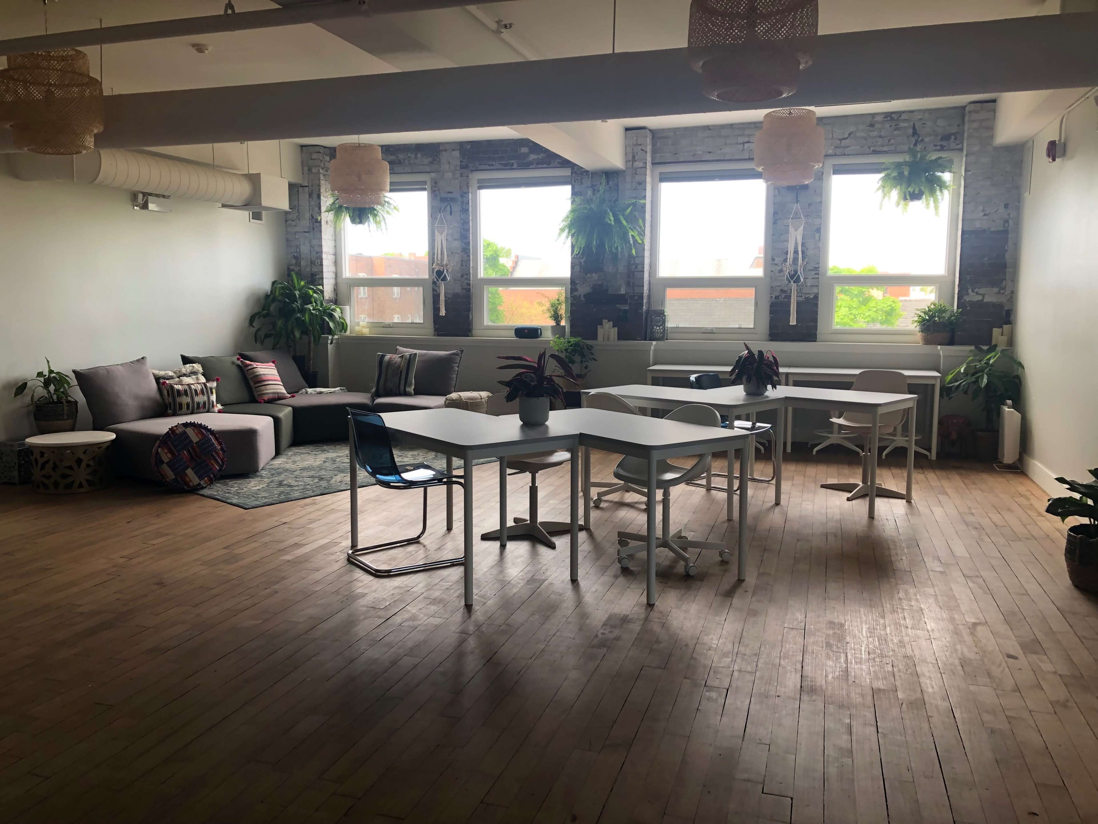 A bright, open office space features large windows, a comfortable seating area with plants, and several tables and chairs arranged for collaboration.