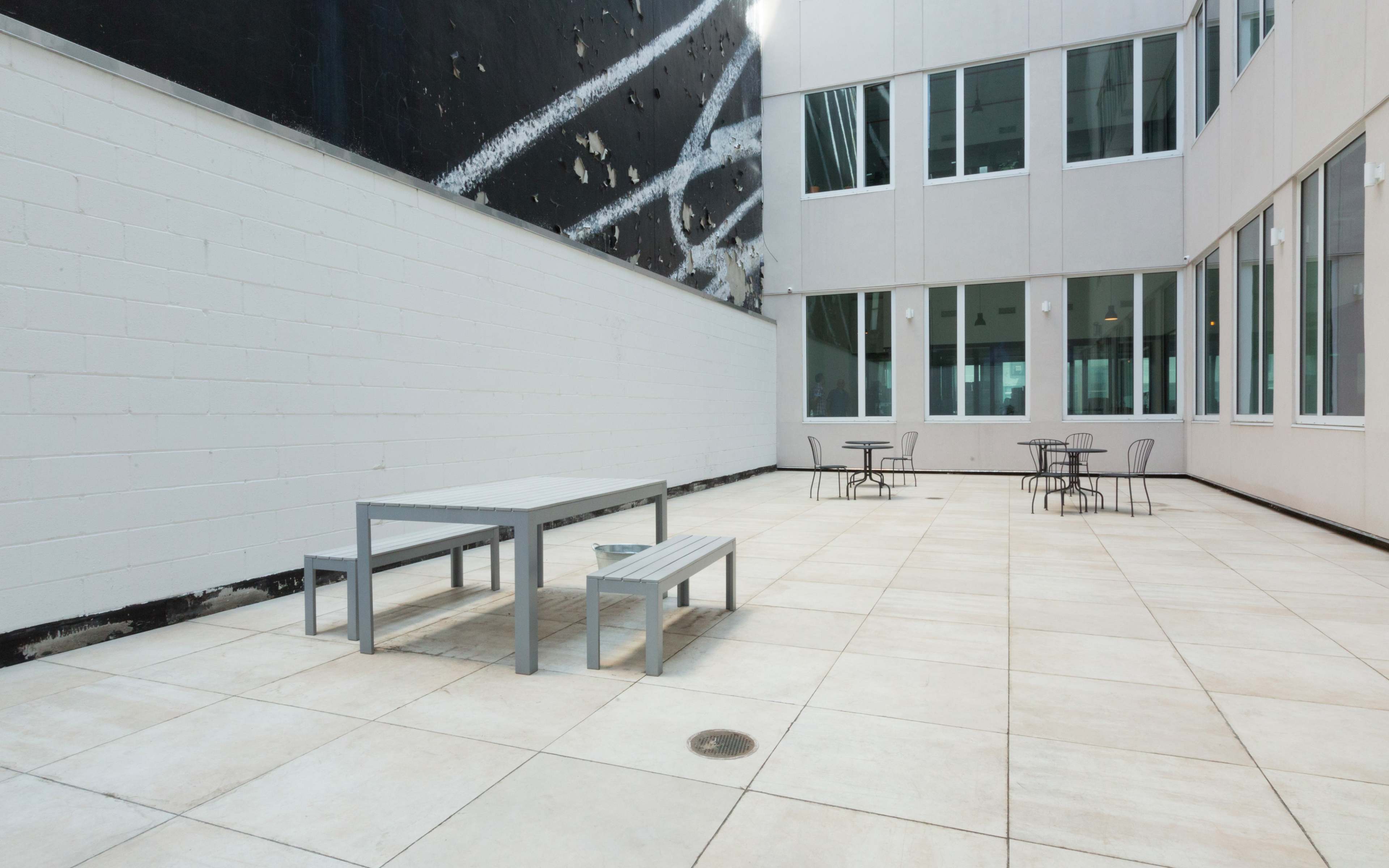 A modern outdoor courtyard with gray tables and benches surrounded by stark white walls and large windows.