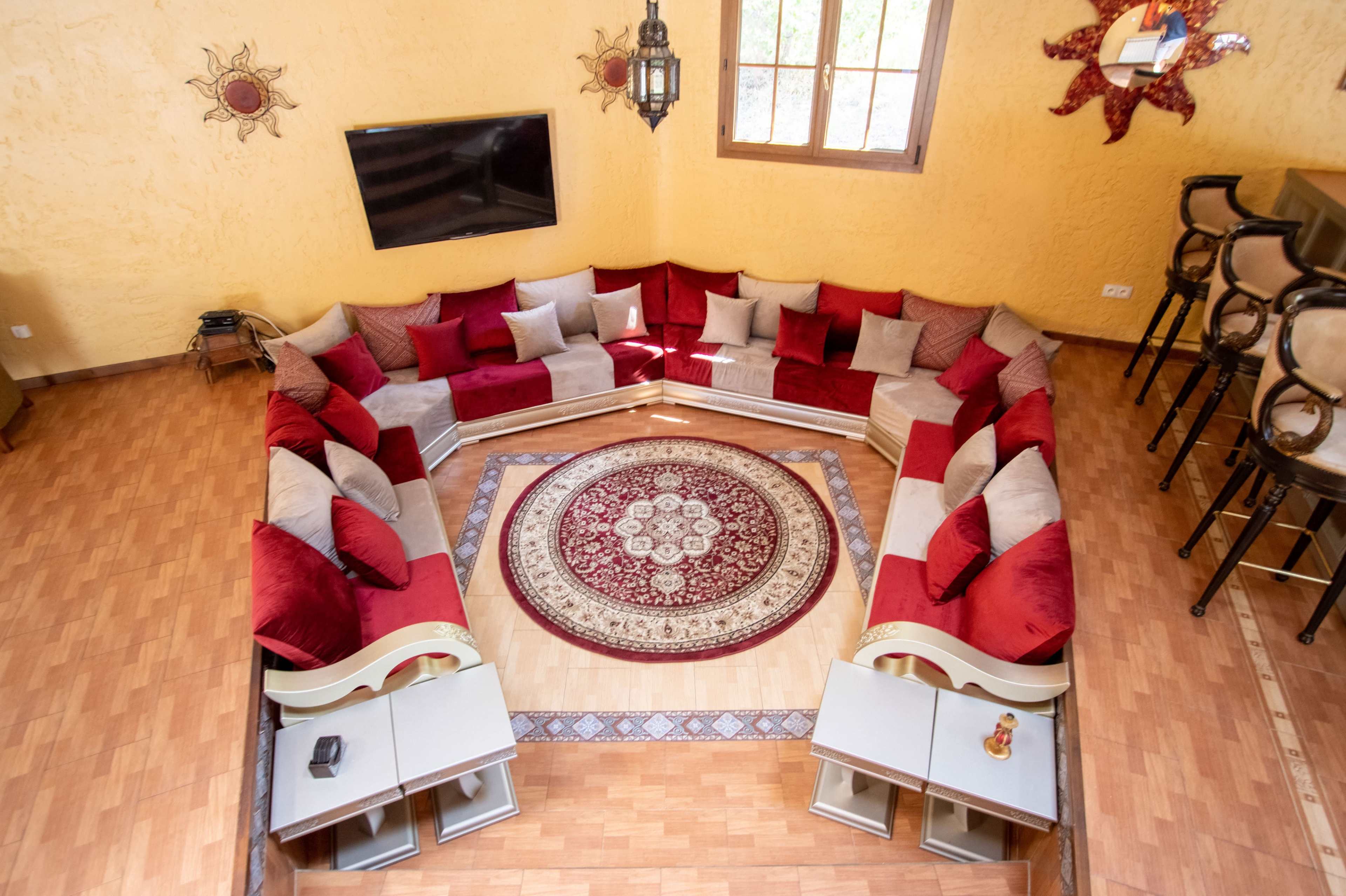 The image shows a spacious living area with a circular arrangement of cushioned seating, a large area rug in the center, a wall-mounted television, and a bar with high stools.