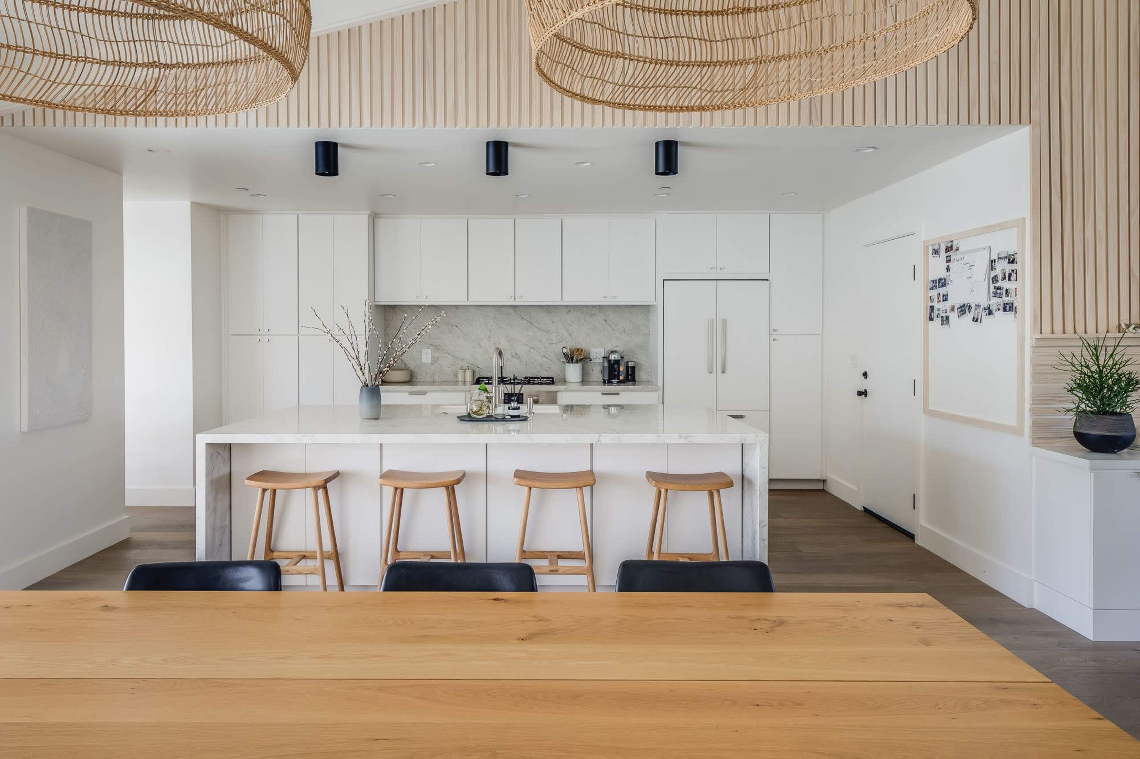 A modern kitchen features a marble island with four wooden stools, white cabinetry, and natural wood accents, adjacent to a dining table.
