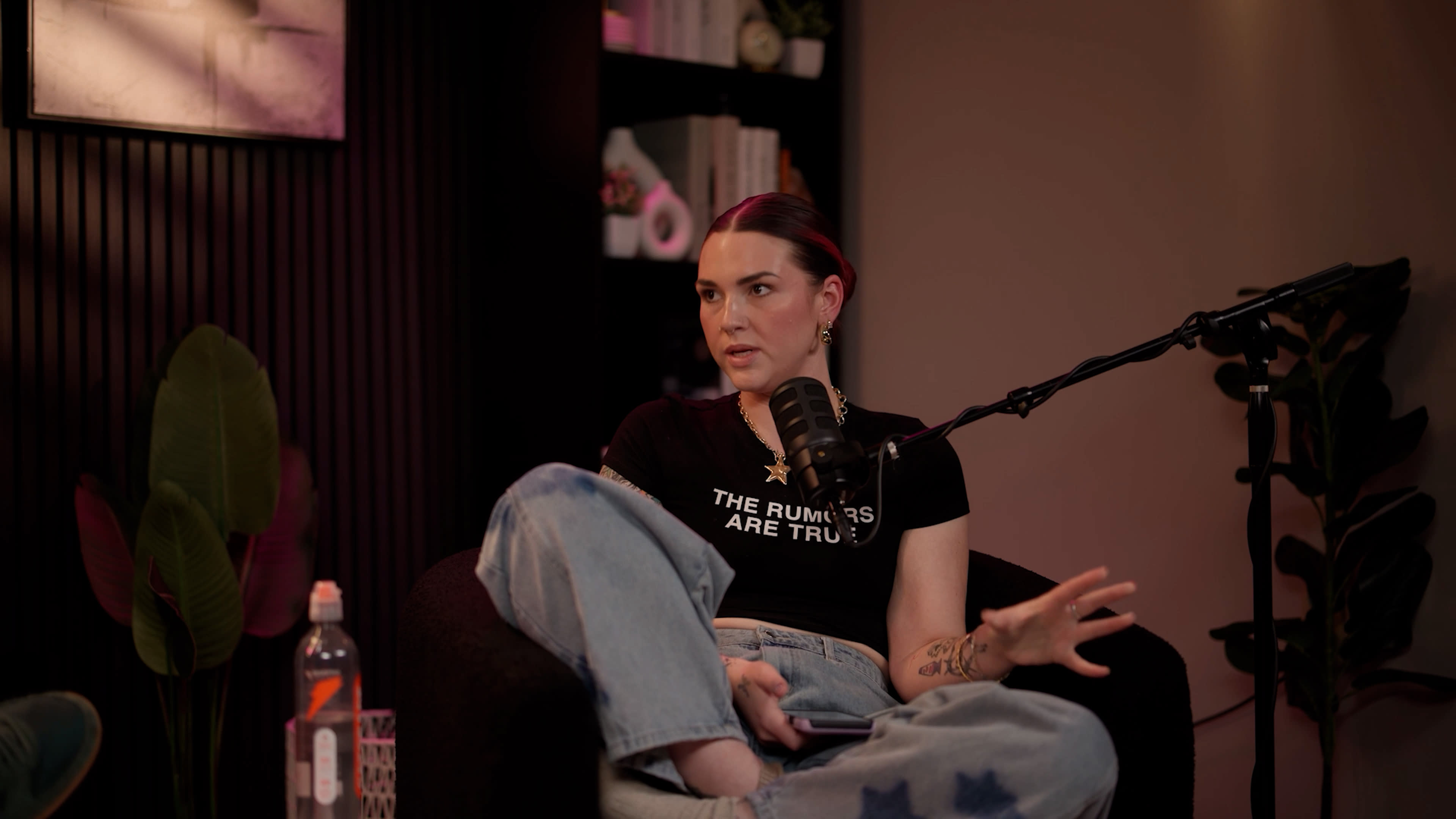 A woman sits on a couch with her legs crossed, speaking into a microphone, while a water bottle and a glass are placed nearby.