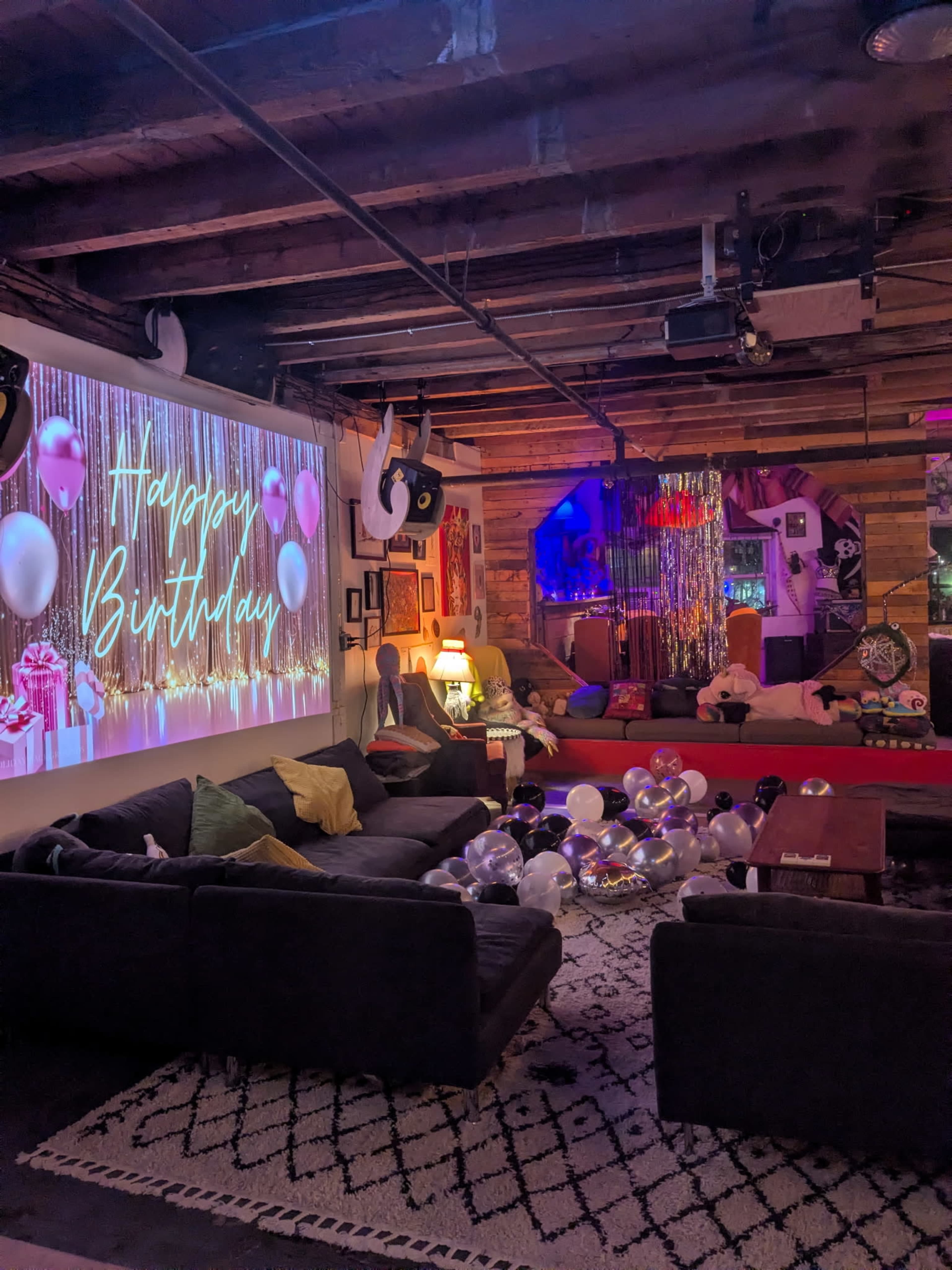 The image shows a festively decorated living space with a "Happy Birthday" projection on the wall, balloons scattered across the floor, and cozy furniture arranged around the room.