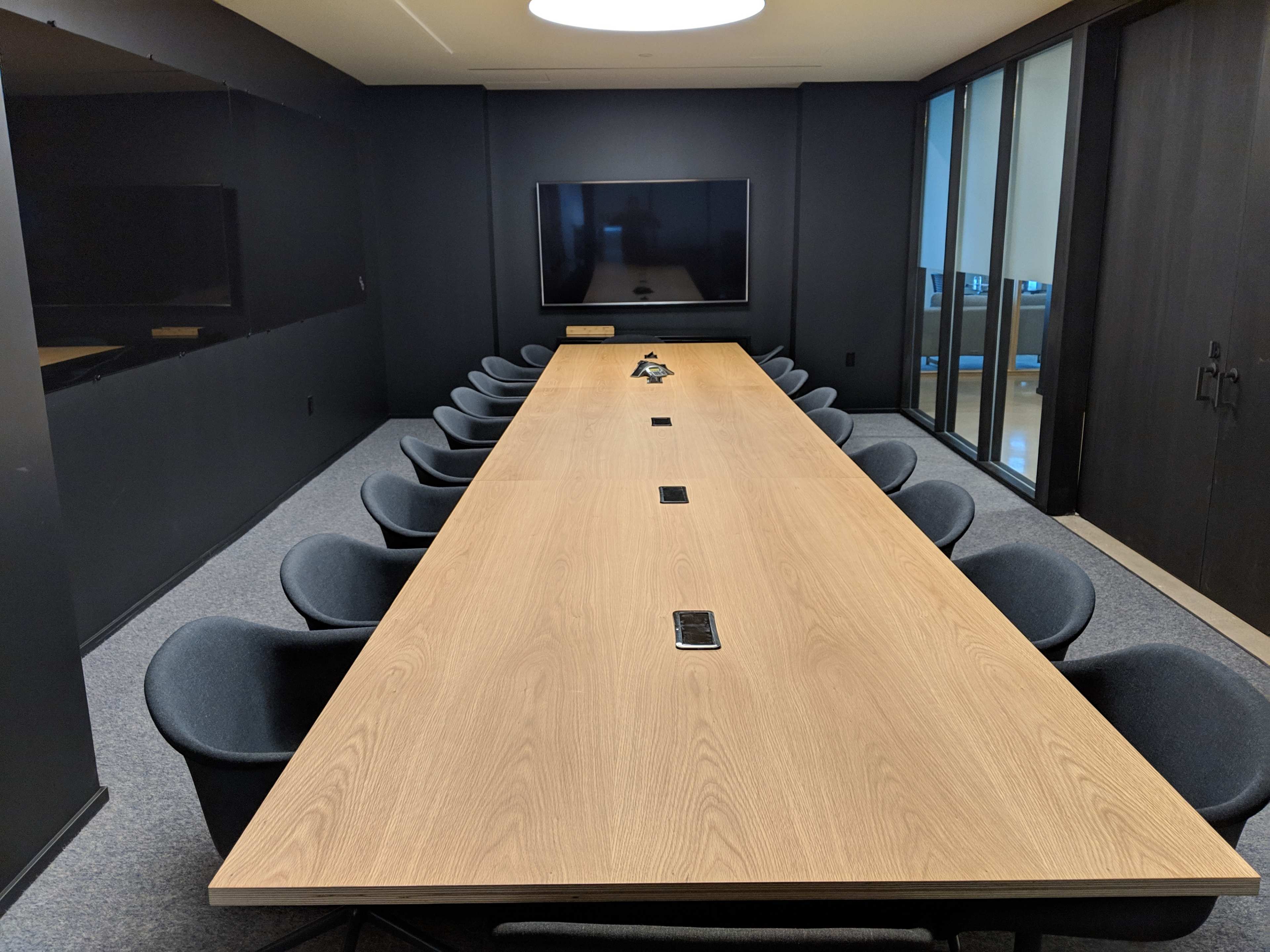 A large conference room with a rectangular wooden table surrounded by black chairs and a television mounted on the wall.