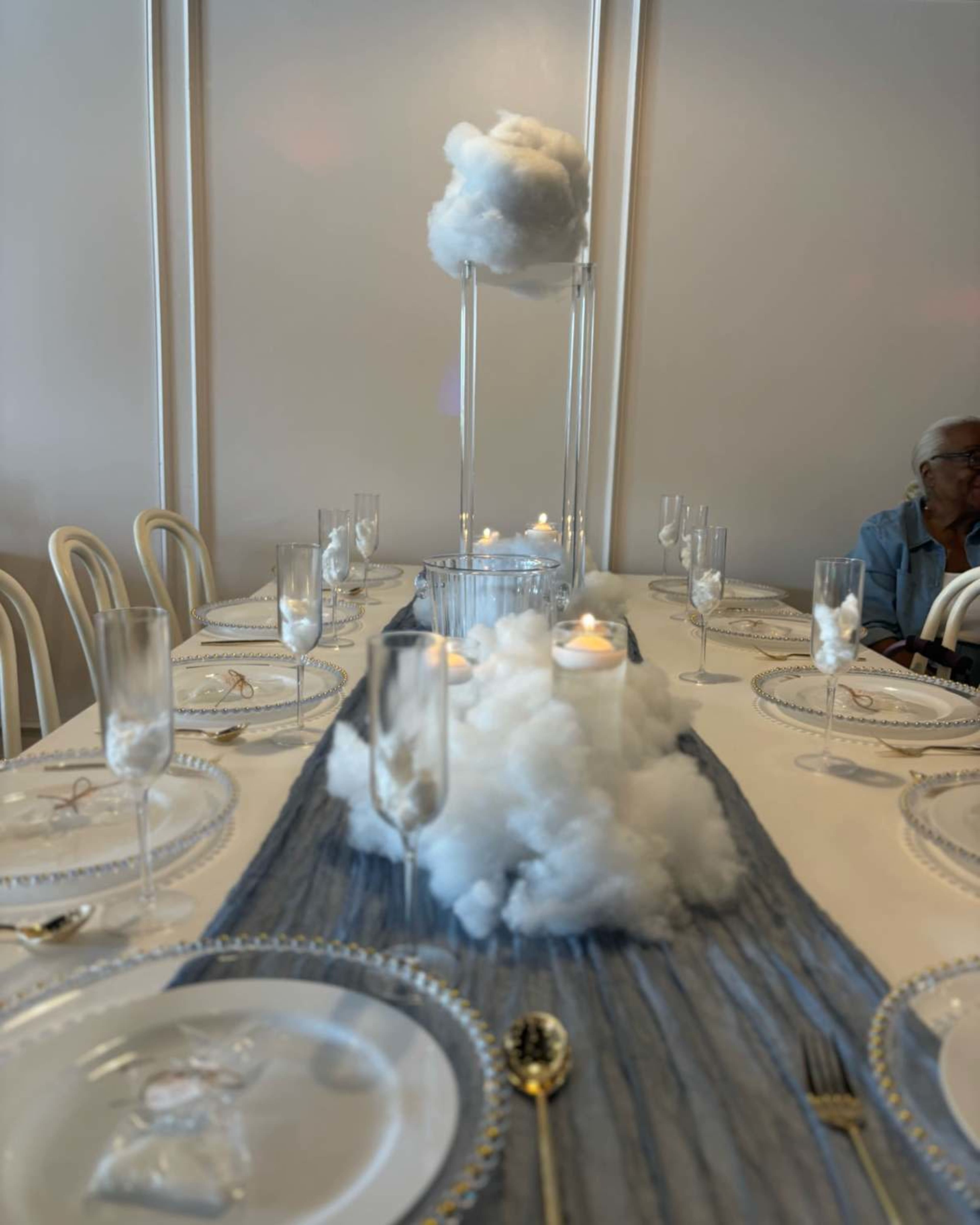 A dining table is set with elegant plates and glasses, featuring decorative cotton clouds and candles as a centerpiece against a light-colored backdrop.