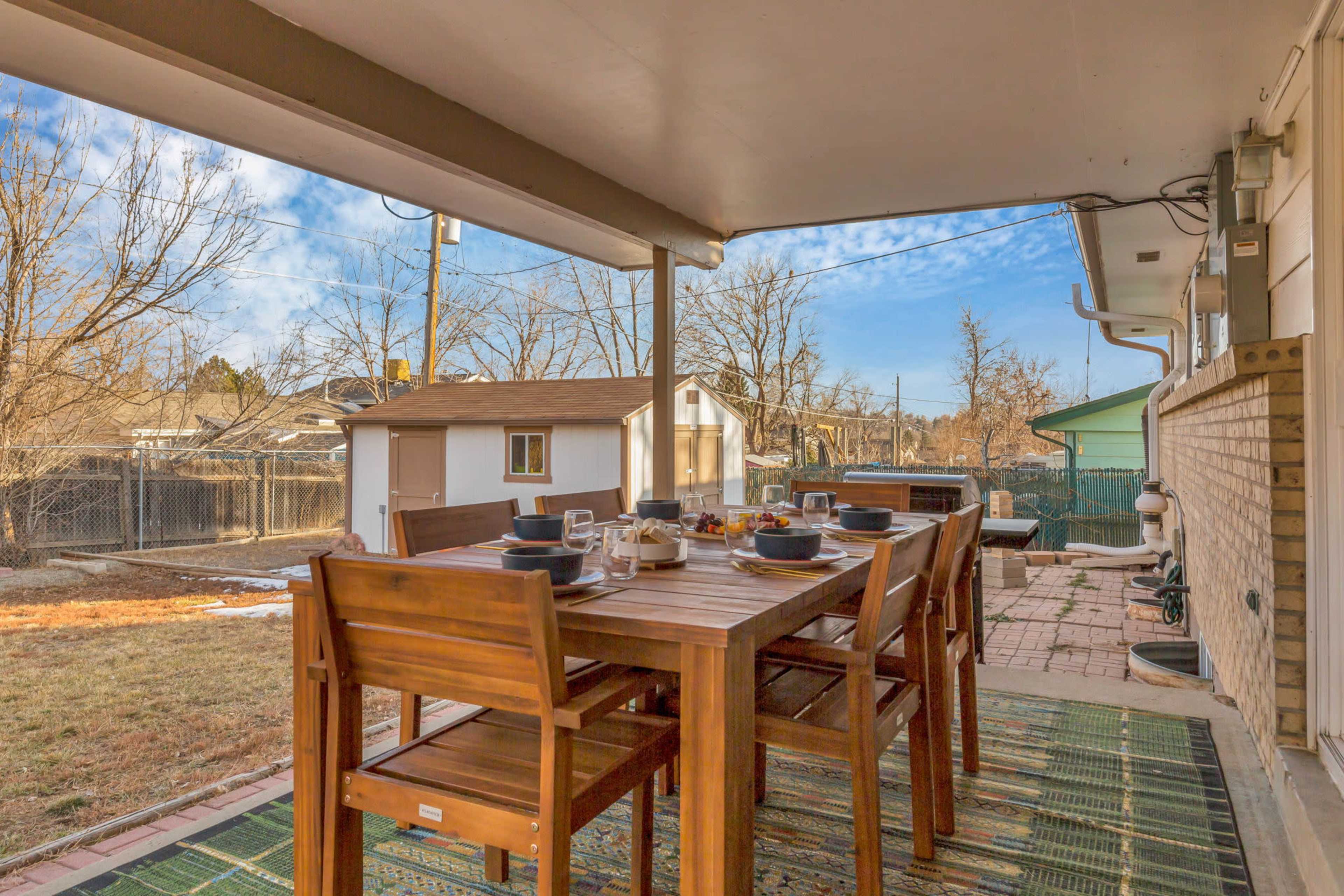 A wooden dining table is set for a meal on a patio with a view of a yard and nearby sheds under a partly cloudy sky.