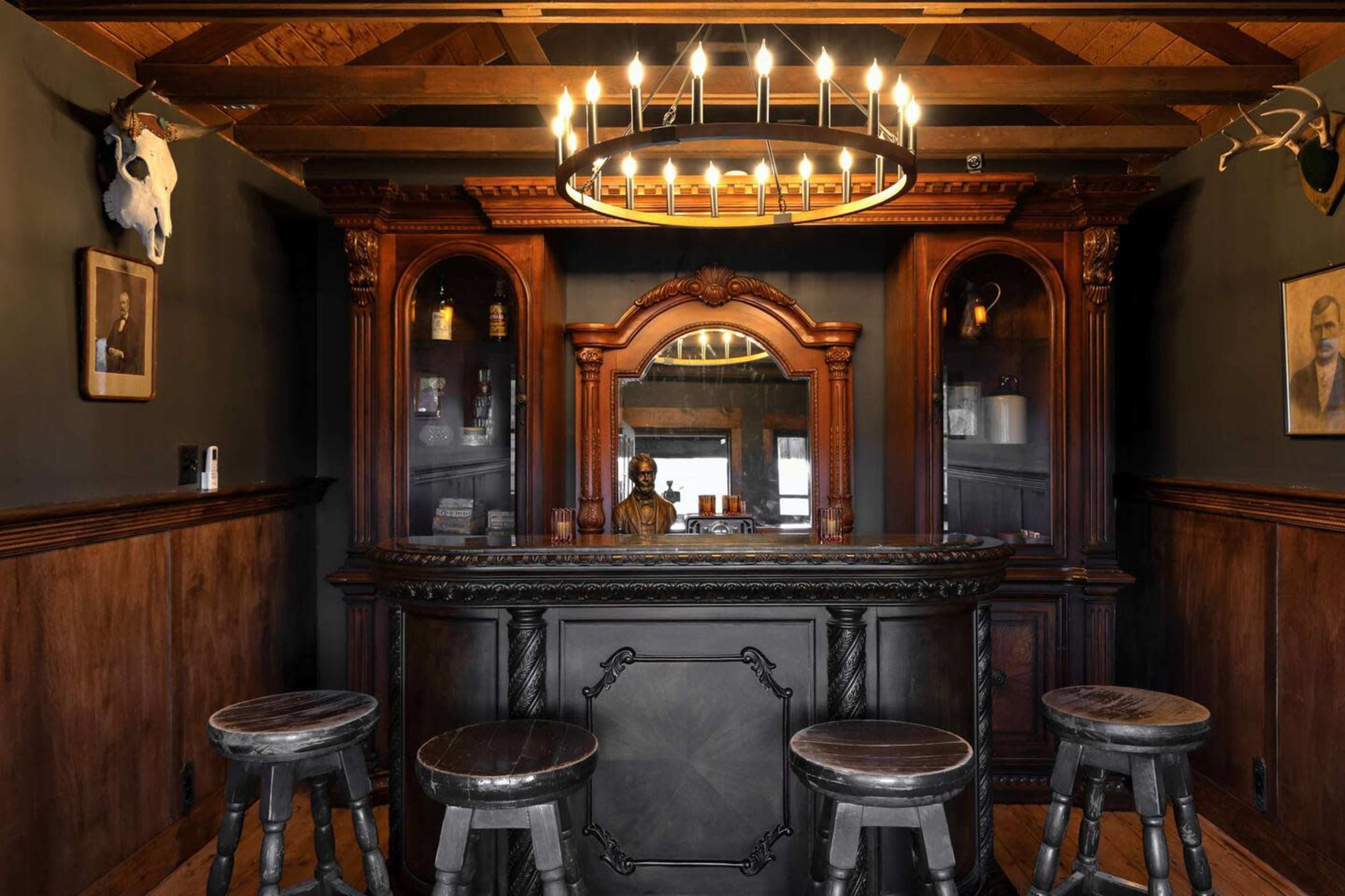 The image shows a wooden bar area with four stools, featuring a vintage-style wooden counter and decorative shelves, framed by dark walls and a chandelier above.