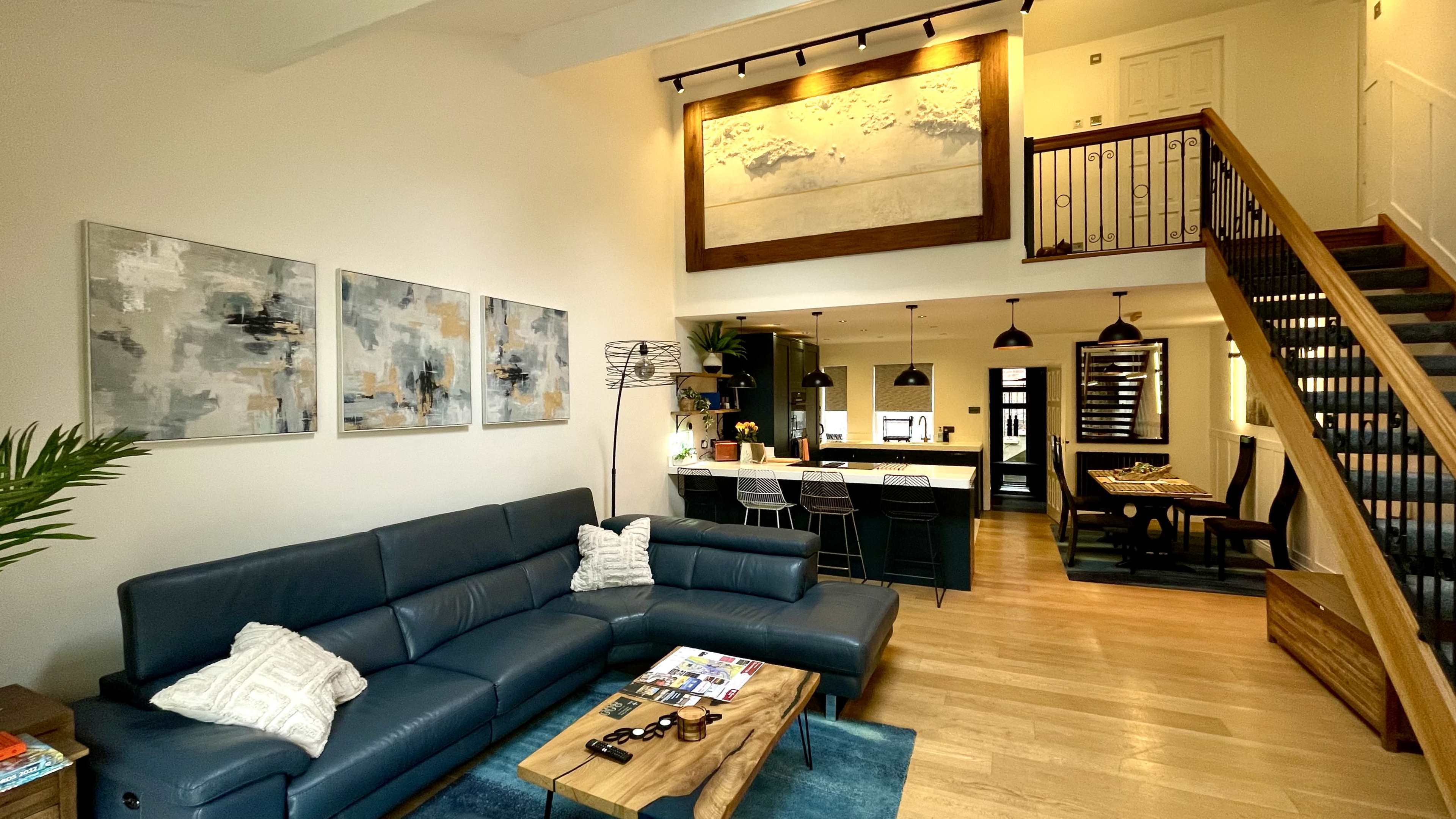The image shows a modern living room with a blue sectional sofa, a wooden coffee table, and artwork on the walls, leading to an open kitchen and a staircase in the background.