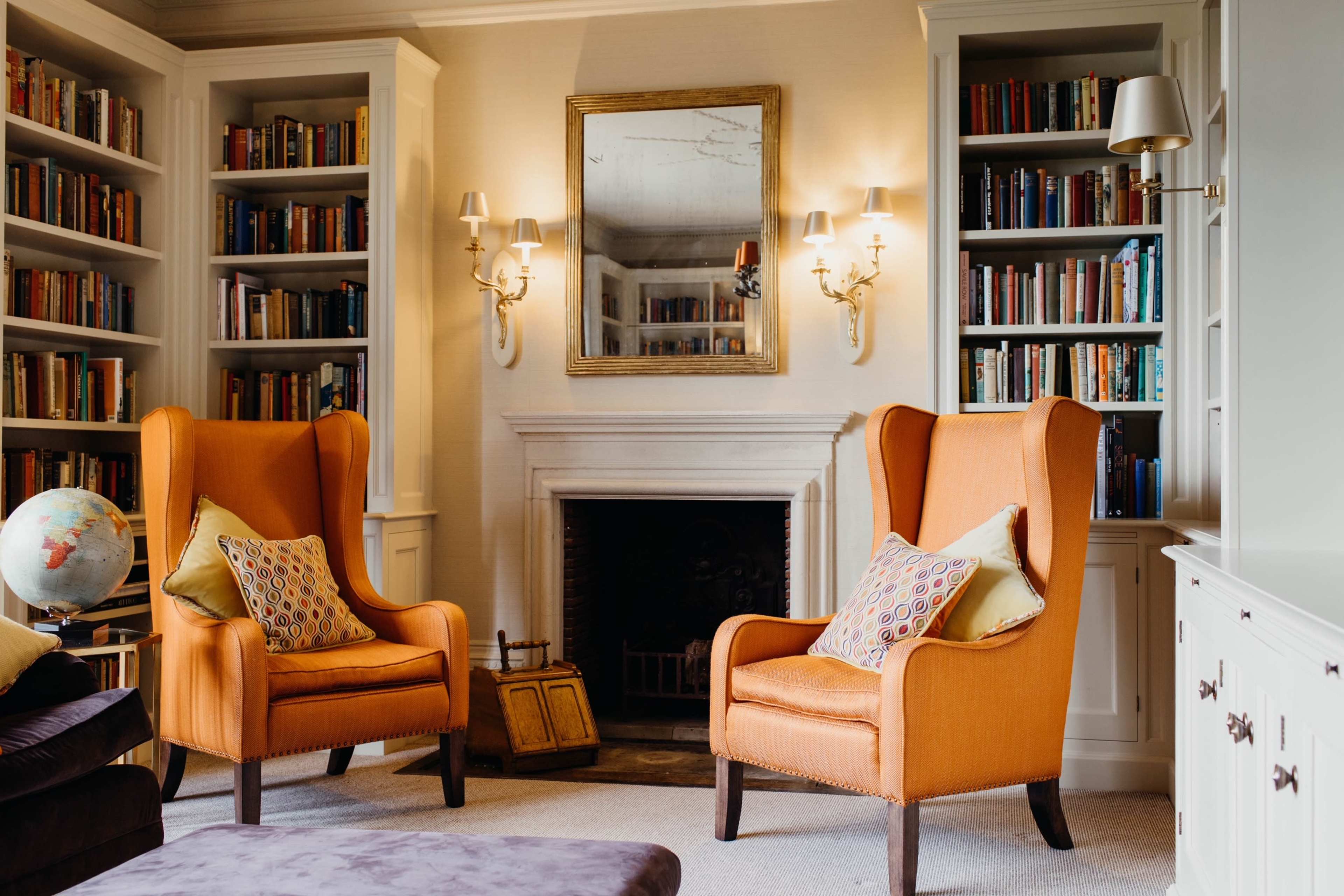 A cozy reading nook with two orange upholstered armchairs, a globe, and bookshelves filled with books, centered around a fireplace.