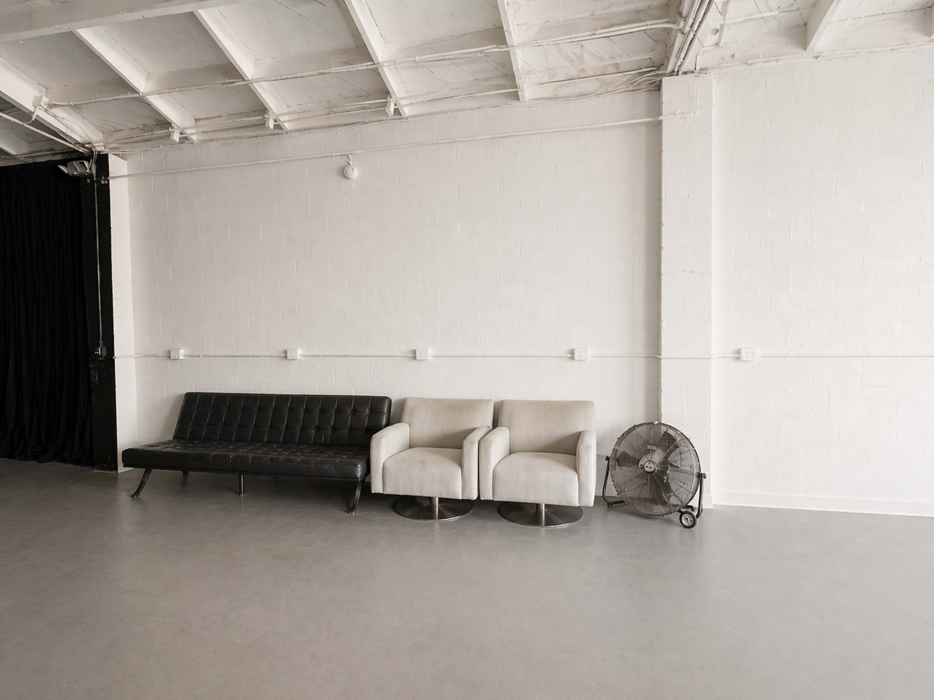 The image shows a minimalist interior space with a black sofa, two light-colored chairs, and a standing fan against a white wall.