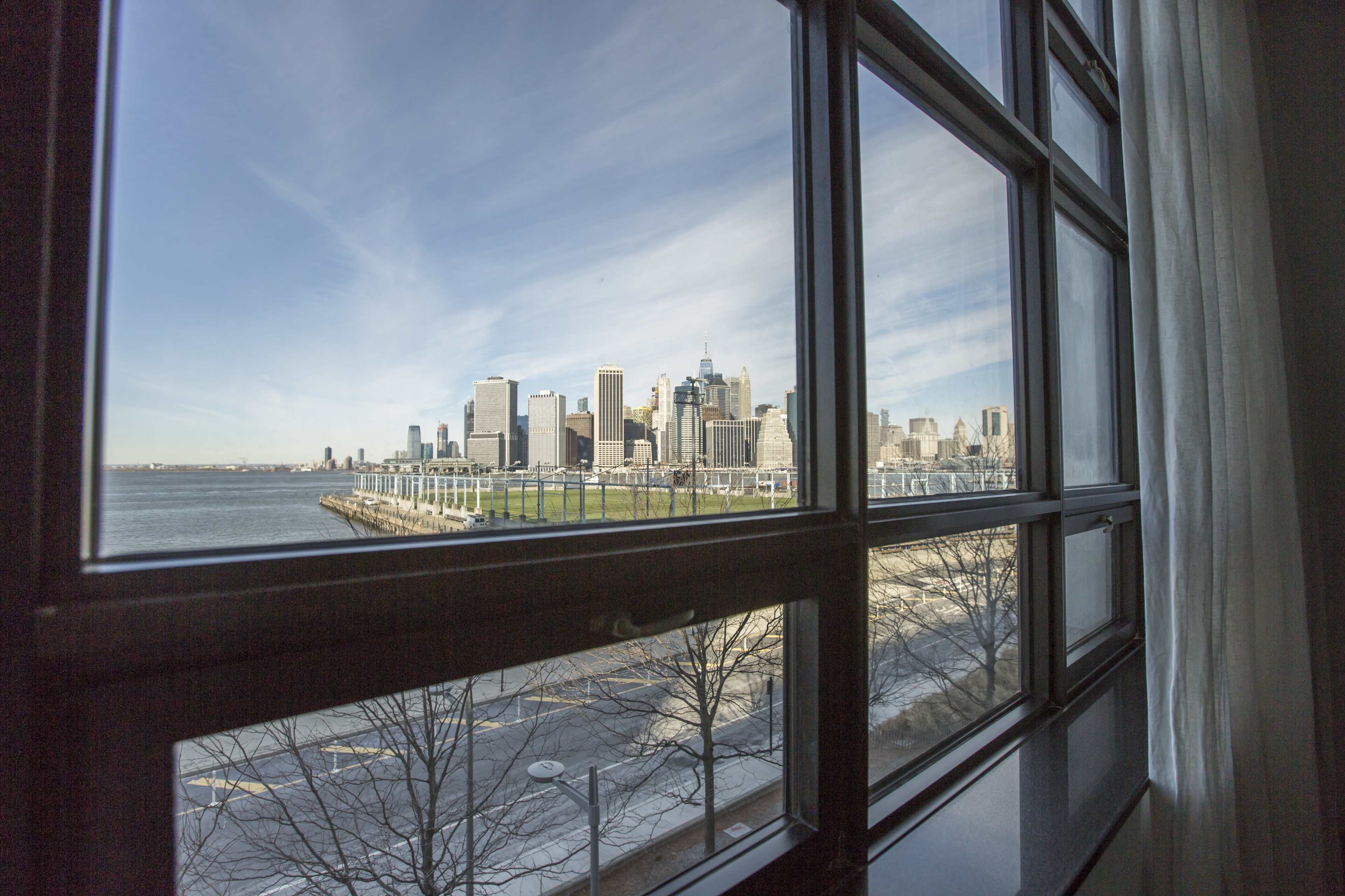 A view of a city skyline across the water, framed by a window with a partially open curtain.