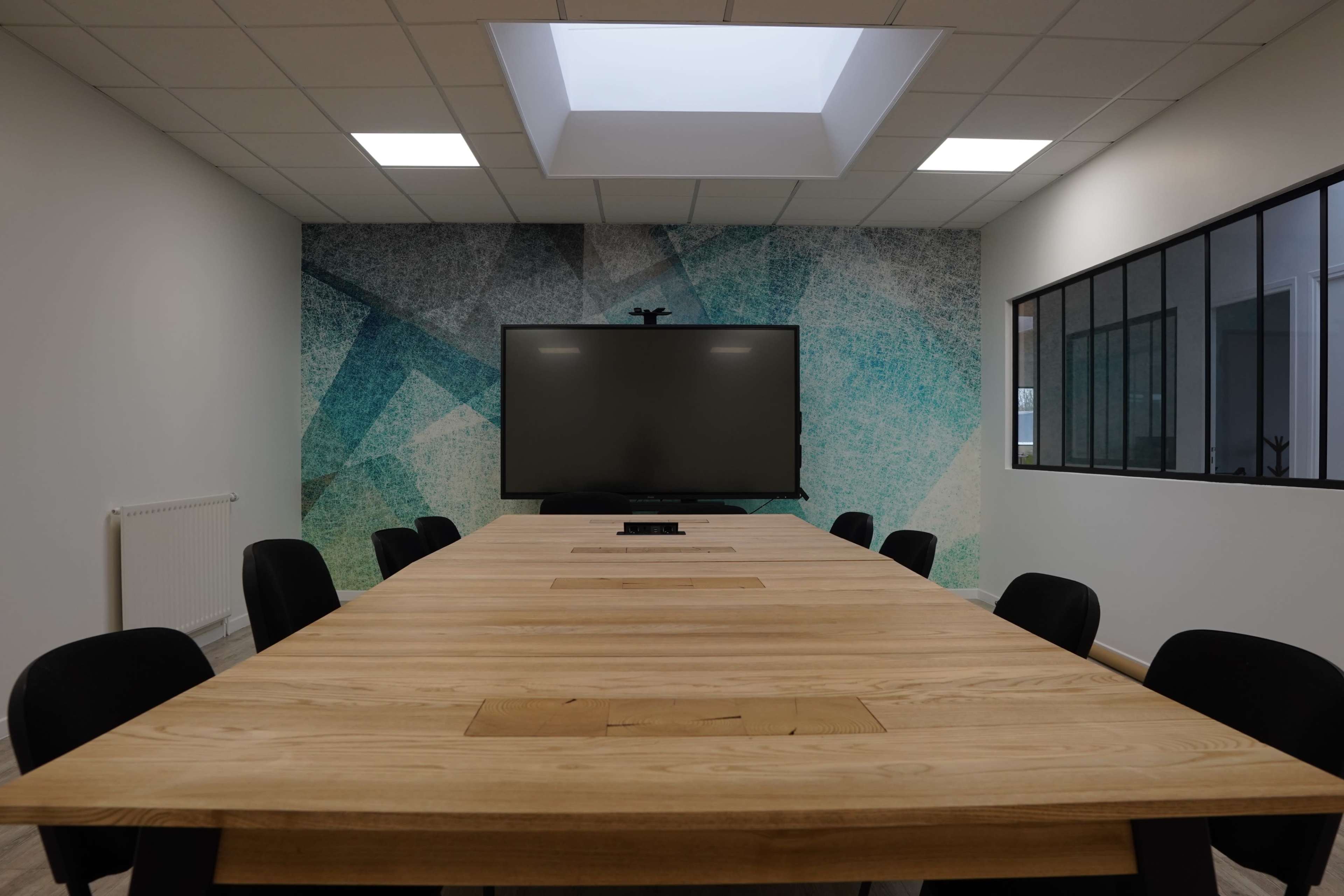 A modern conference room features a large wooden table surrounded by black chairs, with a wall-mounted screen and a skylight above.