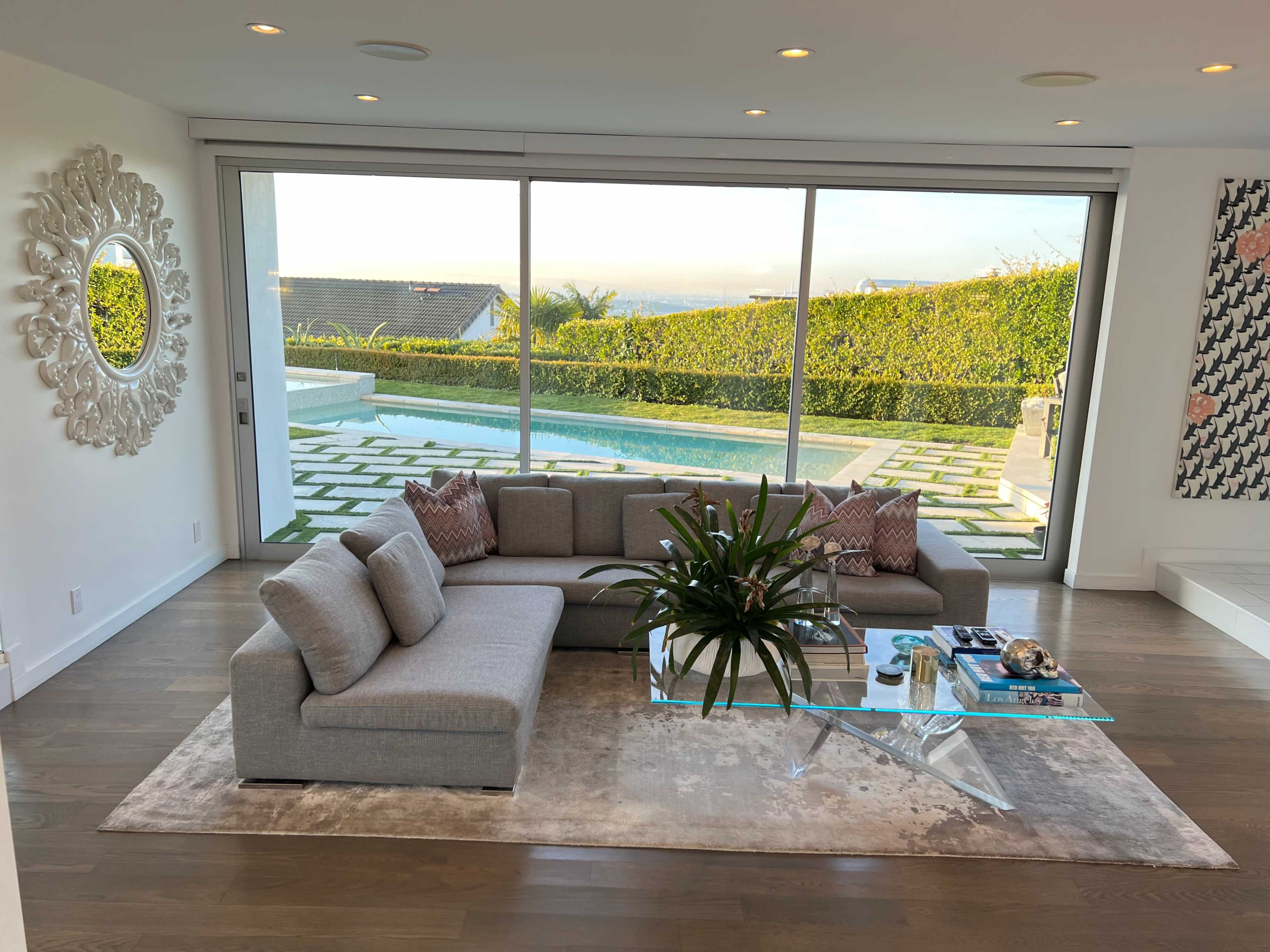 A modern living room with a gray sofa and a glass coffee table, featuring large windows that overlook a pool and landscaped garden.