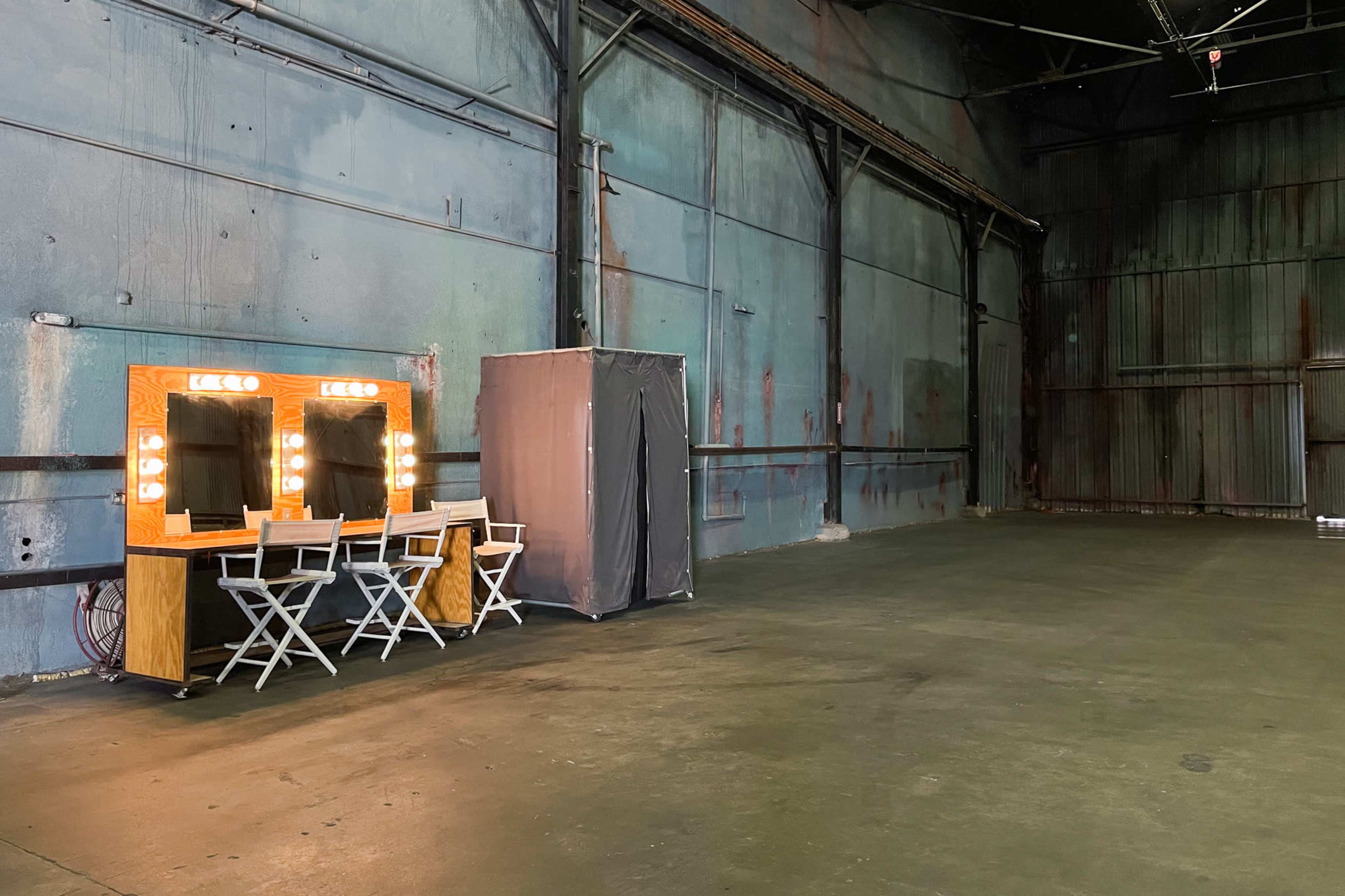 The image shows a spacious, empty warehouse interior with three illuminated vanity mirrors and a privacy screen in one corner.
