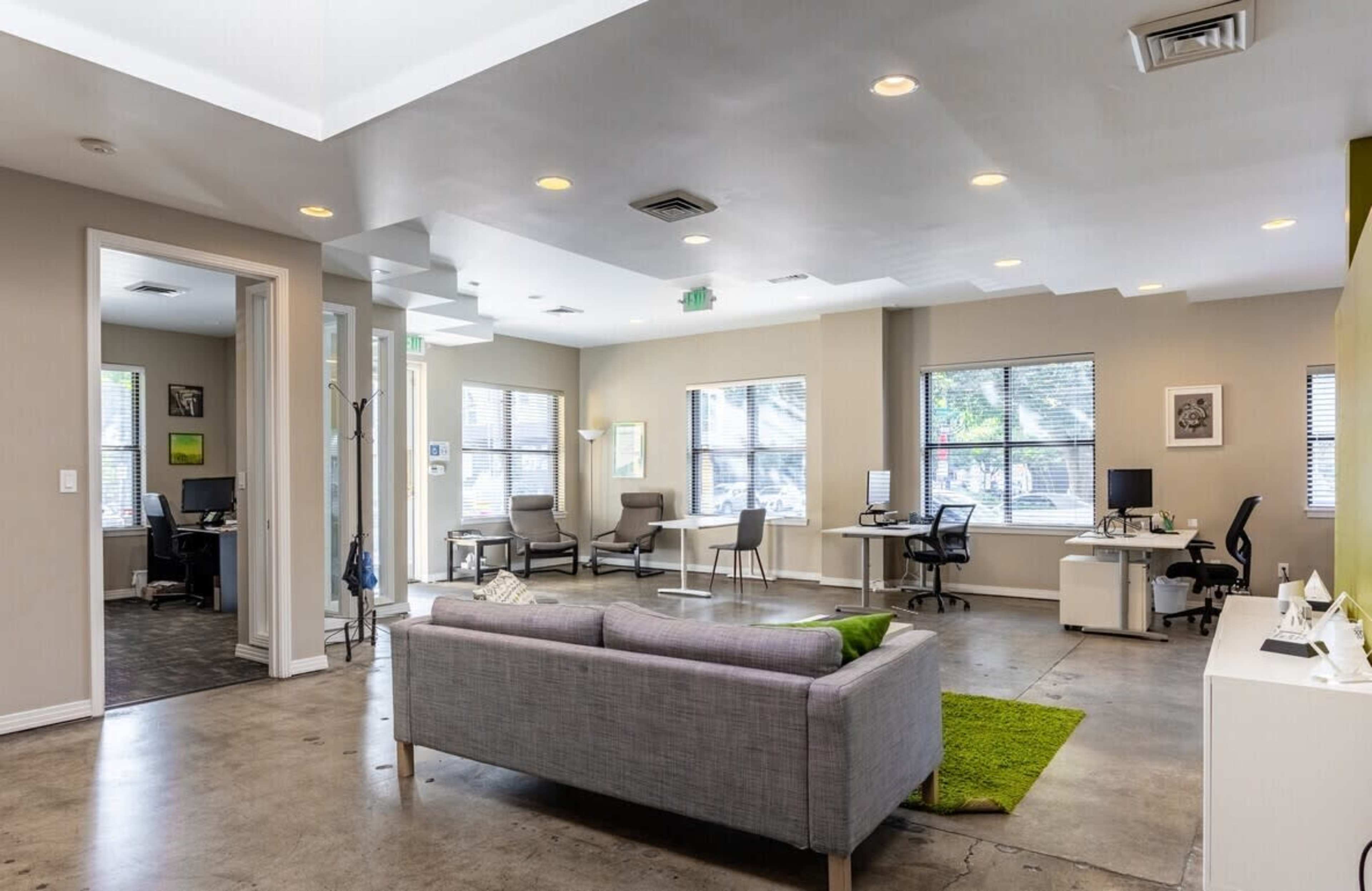 The image shows a modern office space with multiple workstations, a gray couch, and a green area rug on a concrete floor.