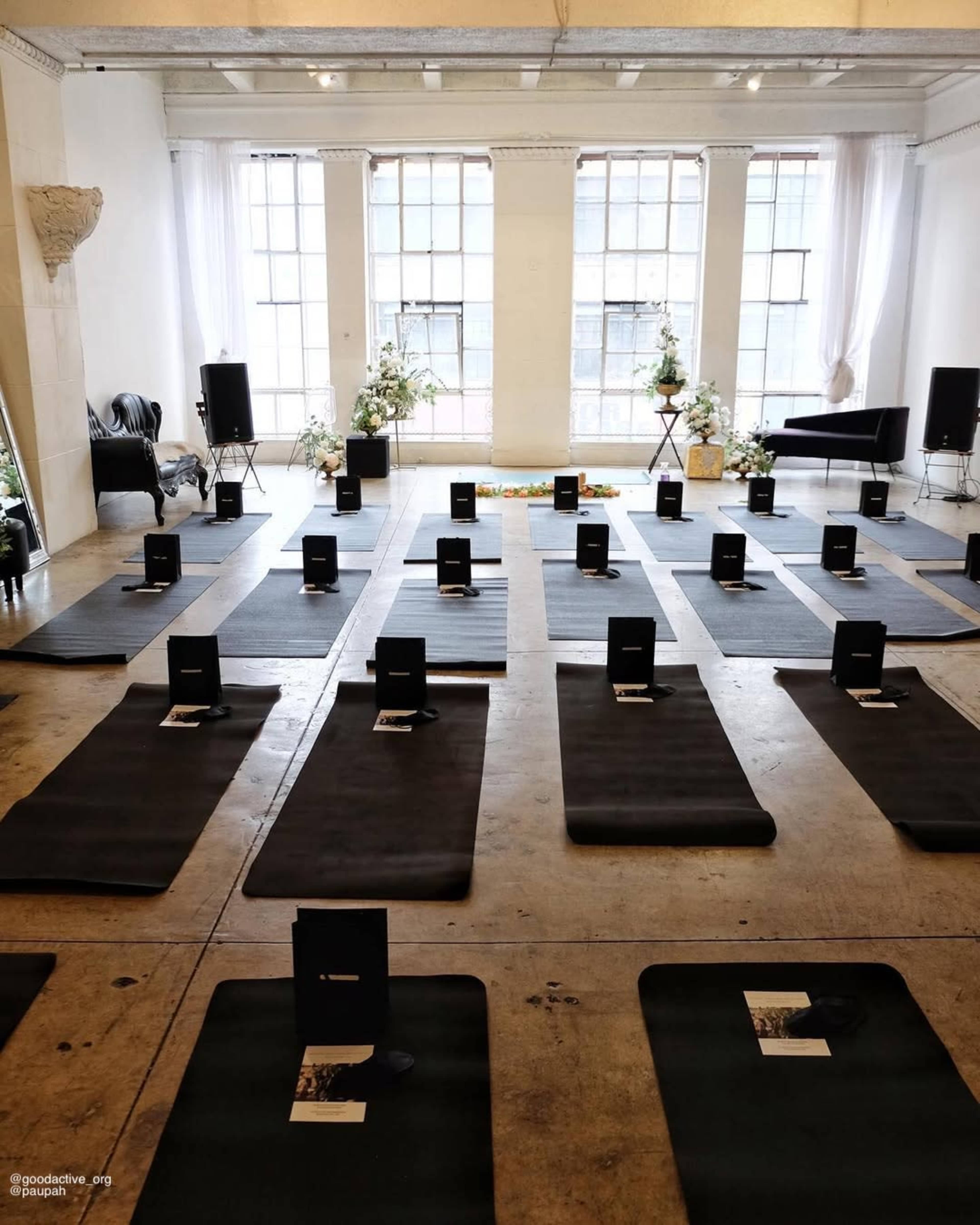 The image shows a spacious room with neatly arranged yoga mats, each featuring a black booklet, and decorated with floral arrangements.