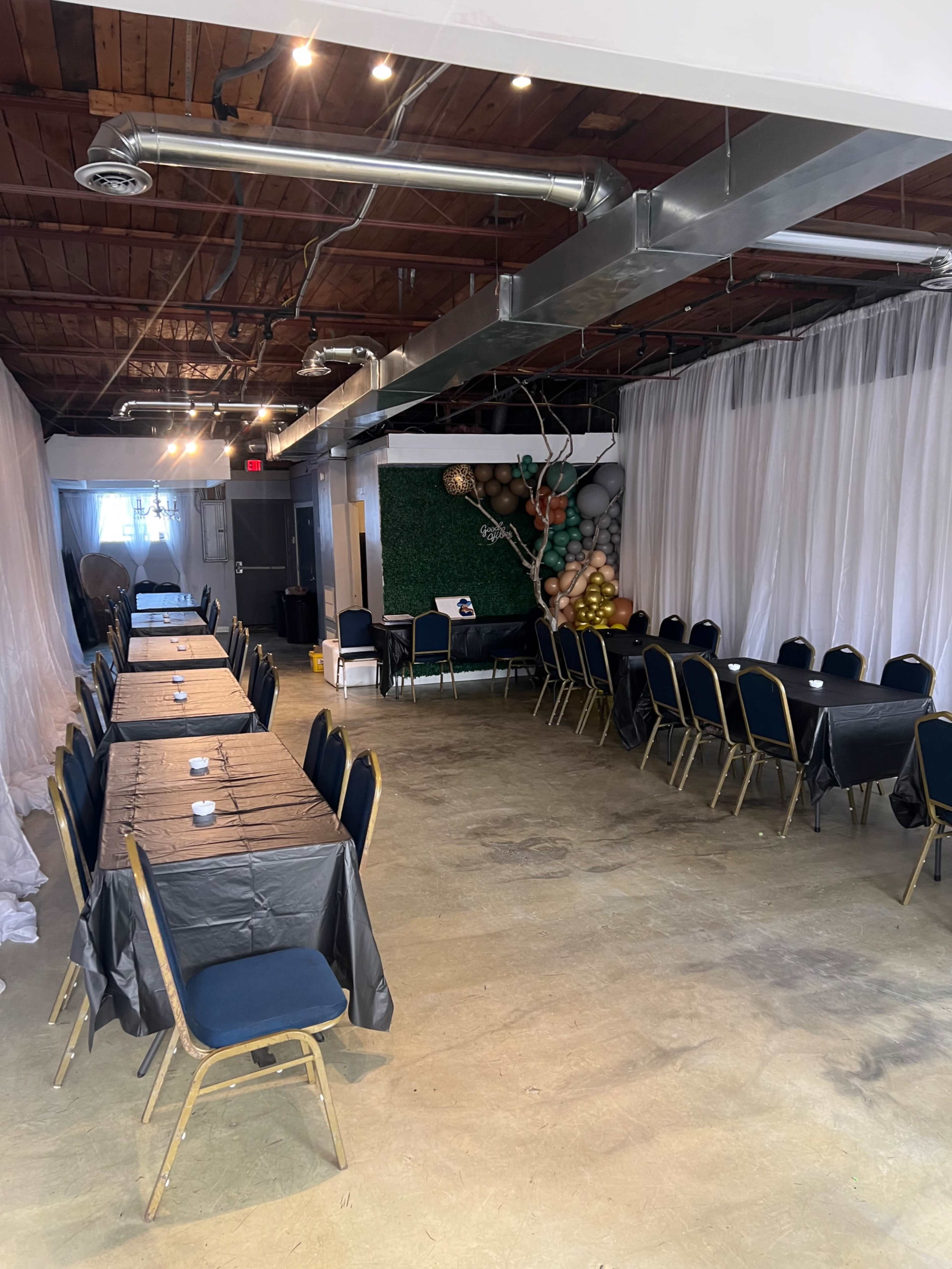 An empty event space with two long tables covered in black cloth, surrounded by blue chairs, and decorative elements including green foliage and balloons in one corner.