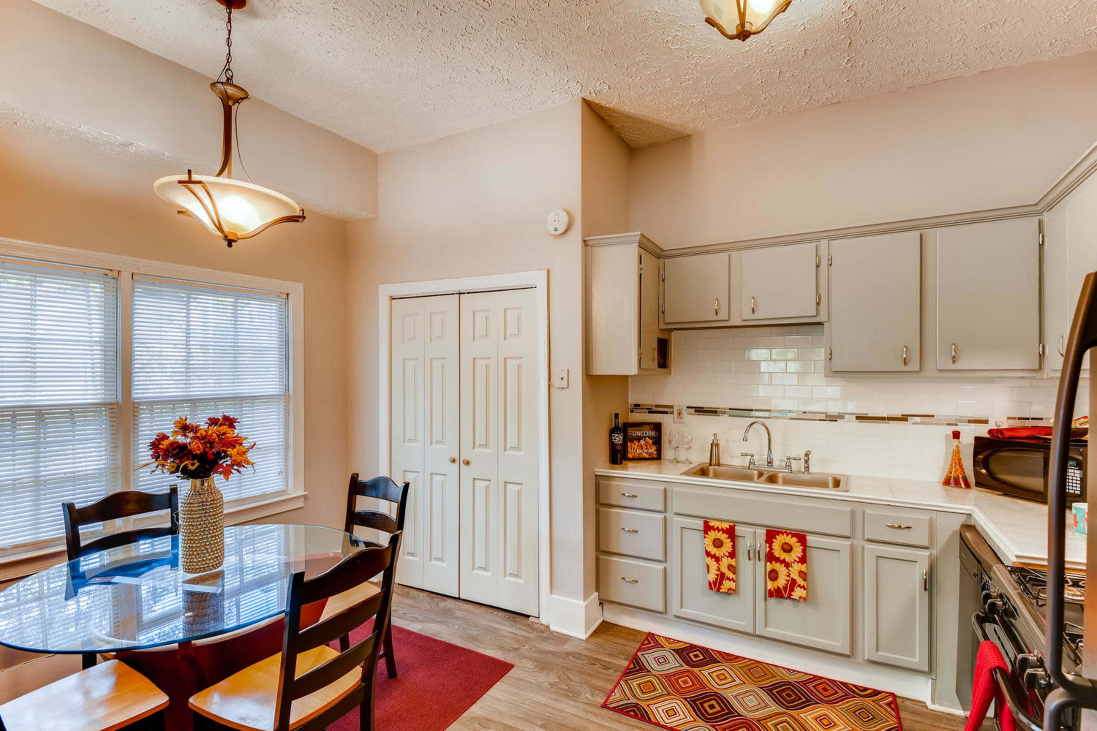A compact kitchen features light gray cabinets, a round dining table with chairs, and a window with natural light.