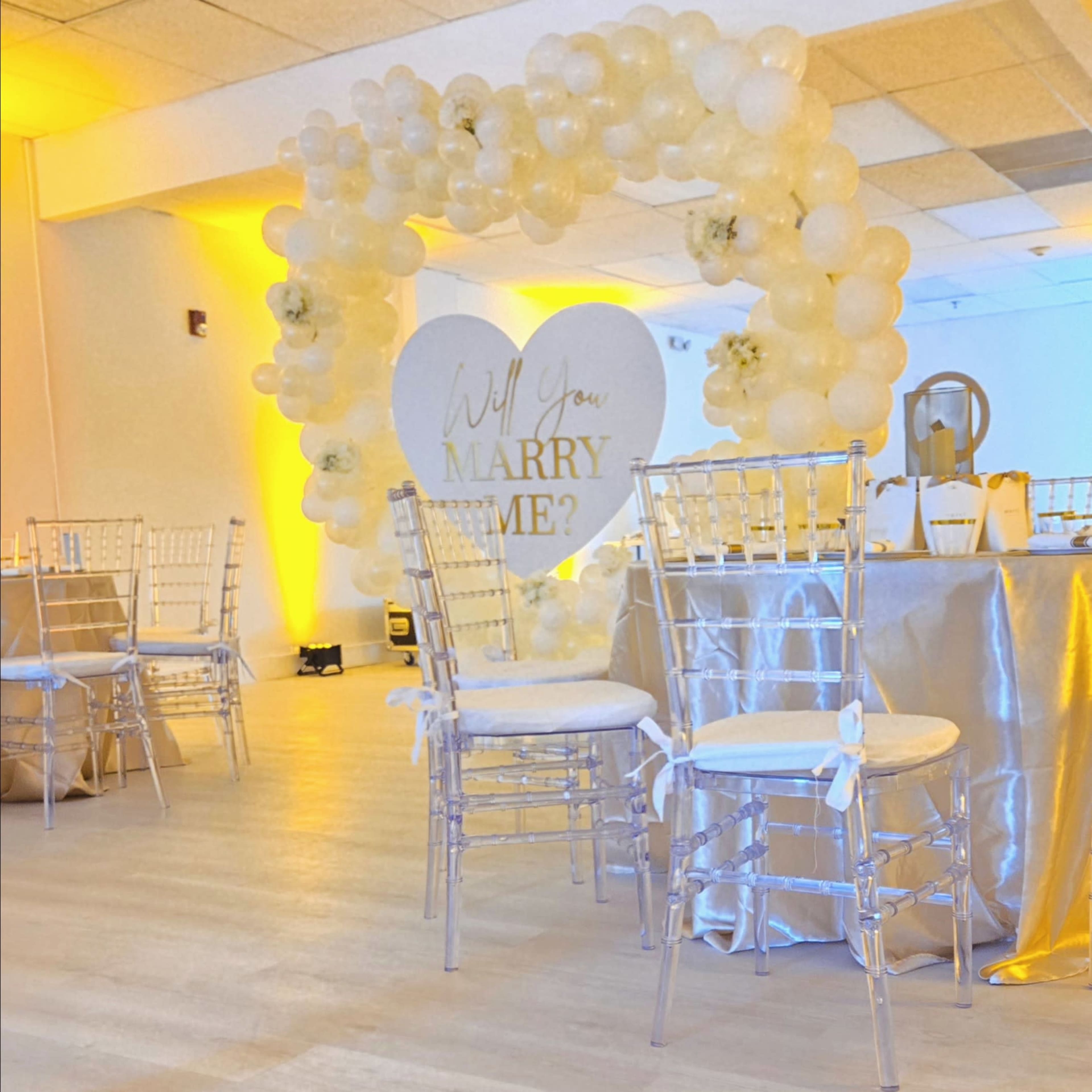 A decorative arch made of balloons surrounds a heart-shaped sign that reads "Will You Marry Me?" in a banquet hall setting with tables and chairs set up.