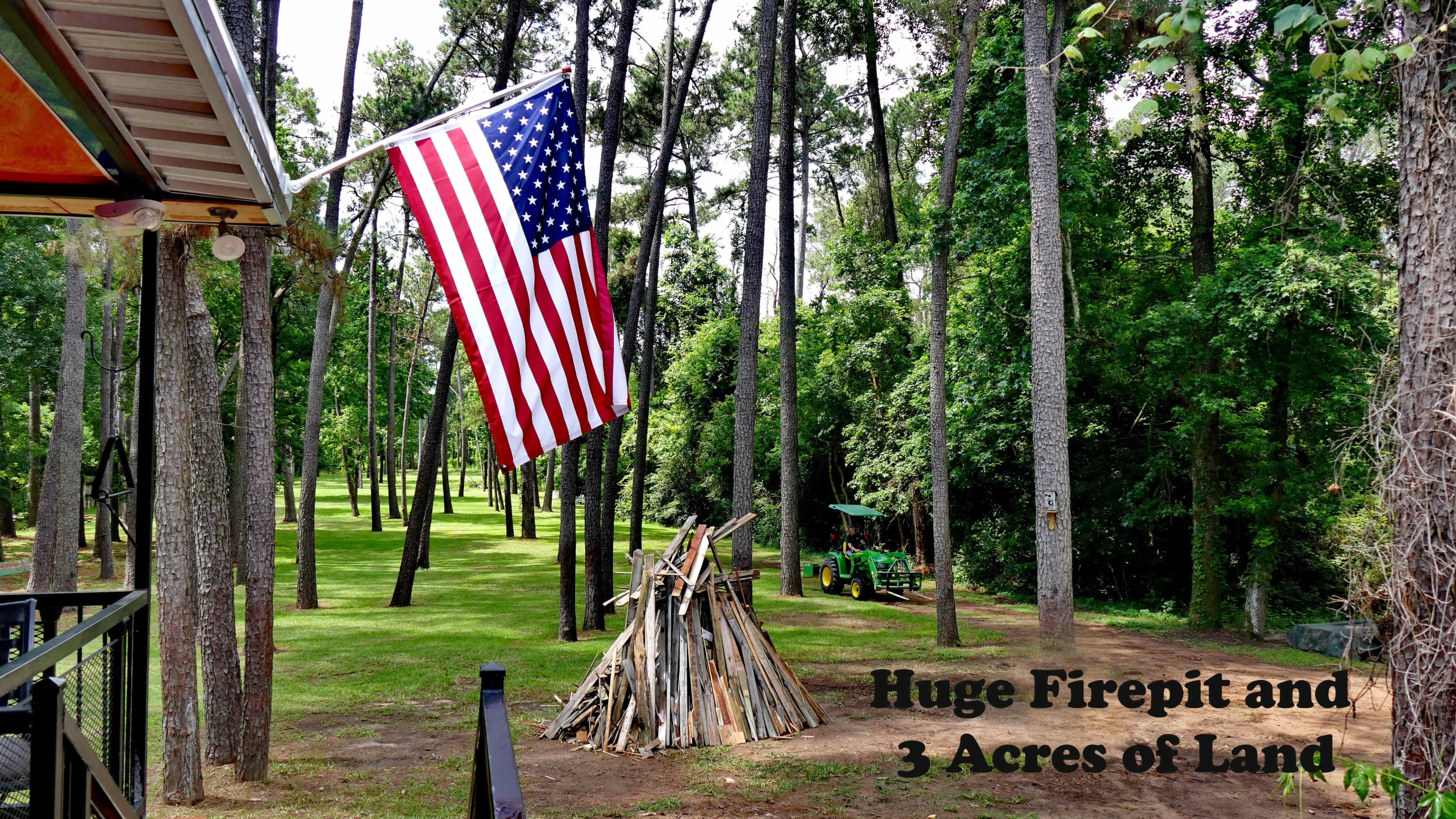 An American flag hanging from a porch, with a large firepit and a pile of firewood in a wooded backyard that spans three acres.