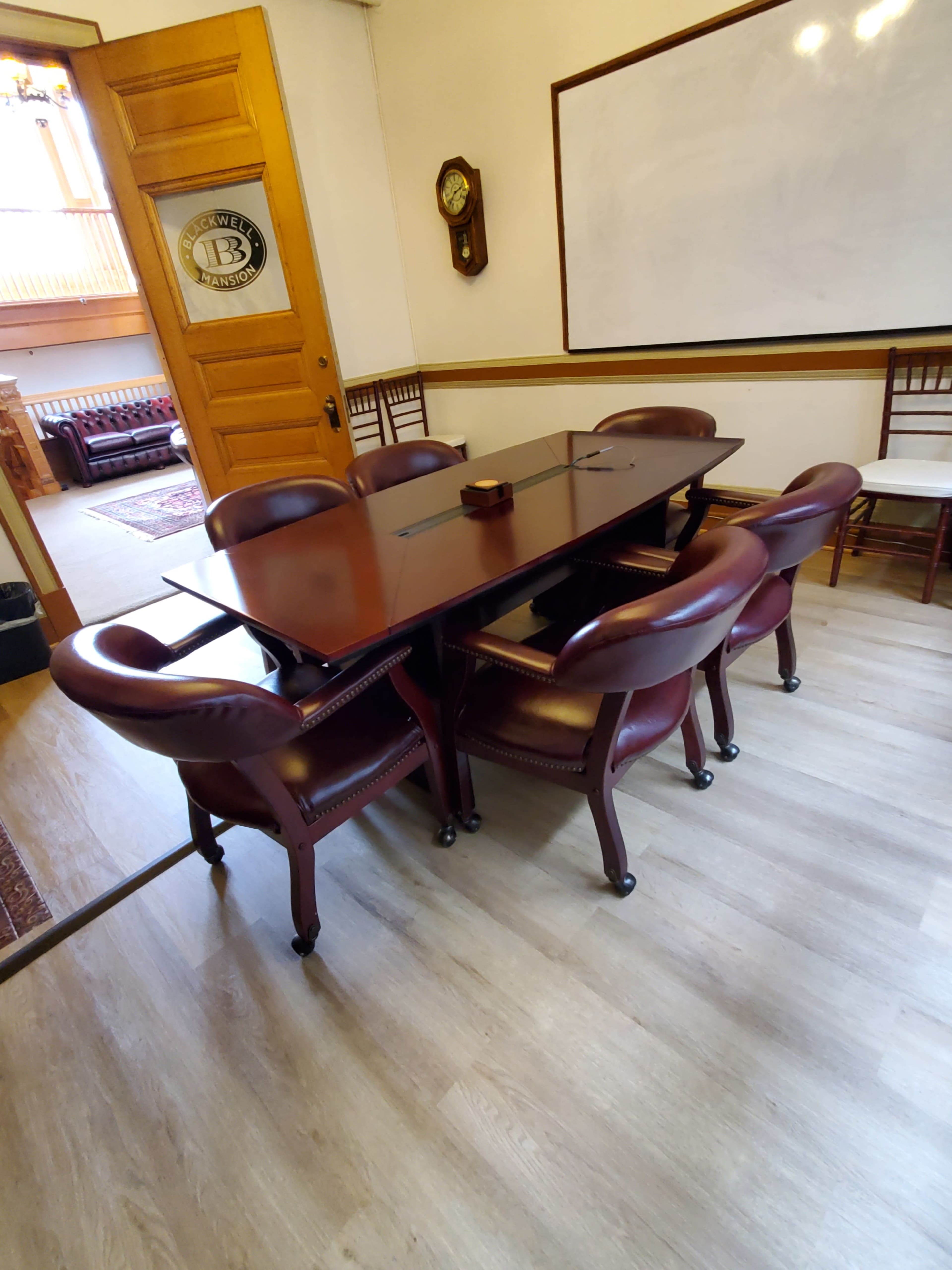 A wooden conference table surrounded by eight leather seating chairs is placed in a brightly lit meeting room.