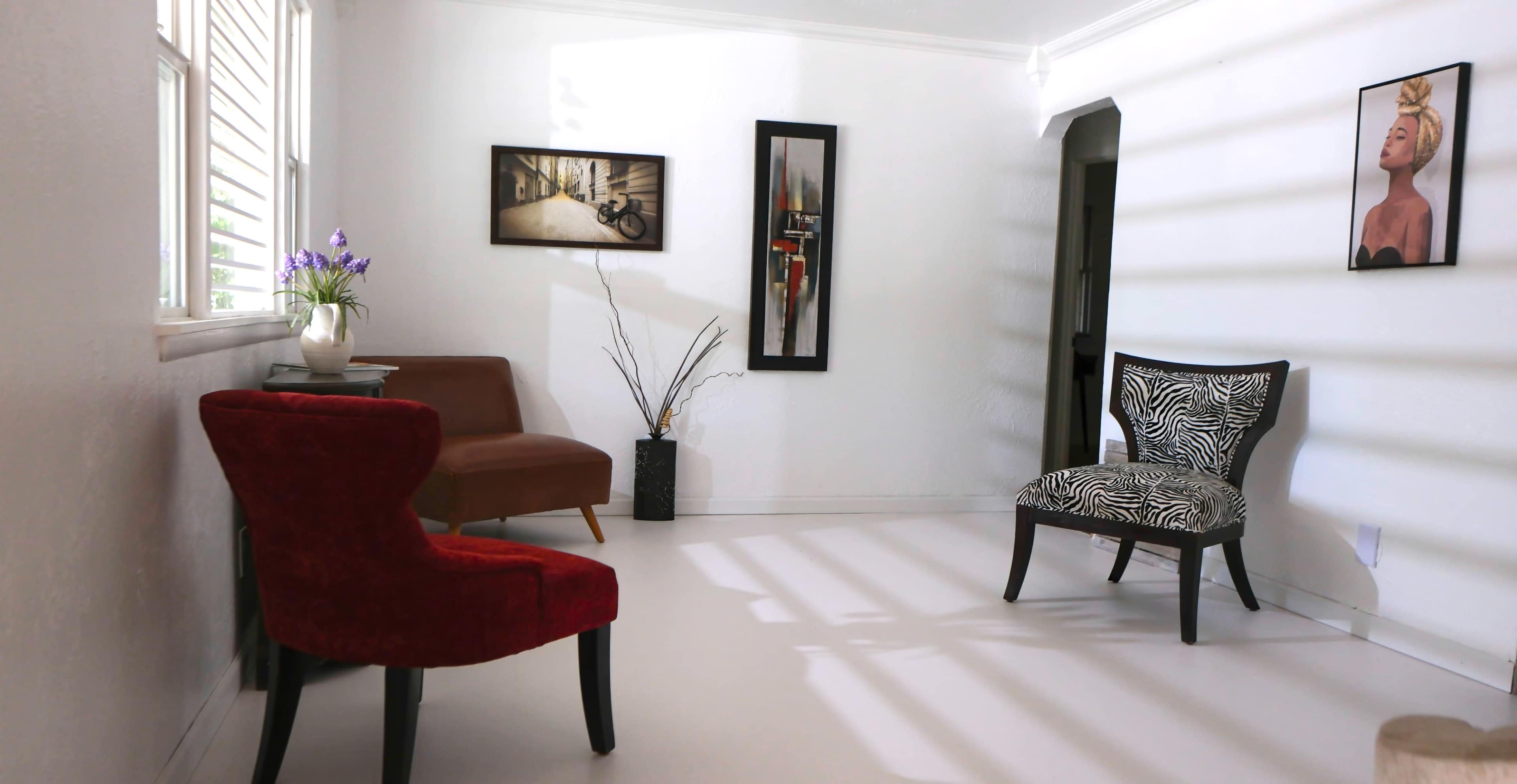 The scene features a brightly lit room with two chairs, one red and one with a zebra print, a small table with a vase of flowers, and two framed artworks on the walls.