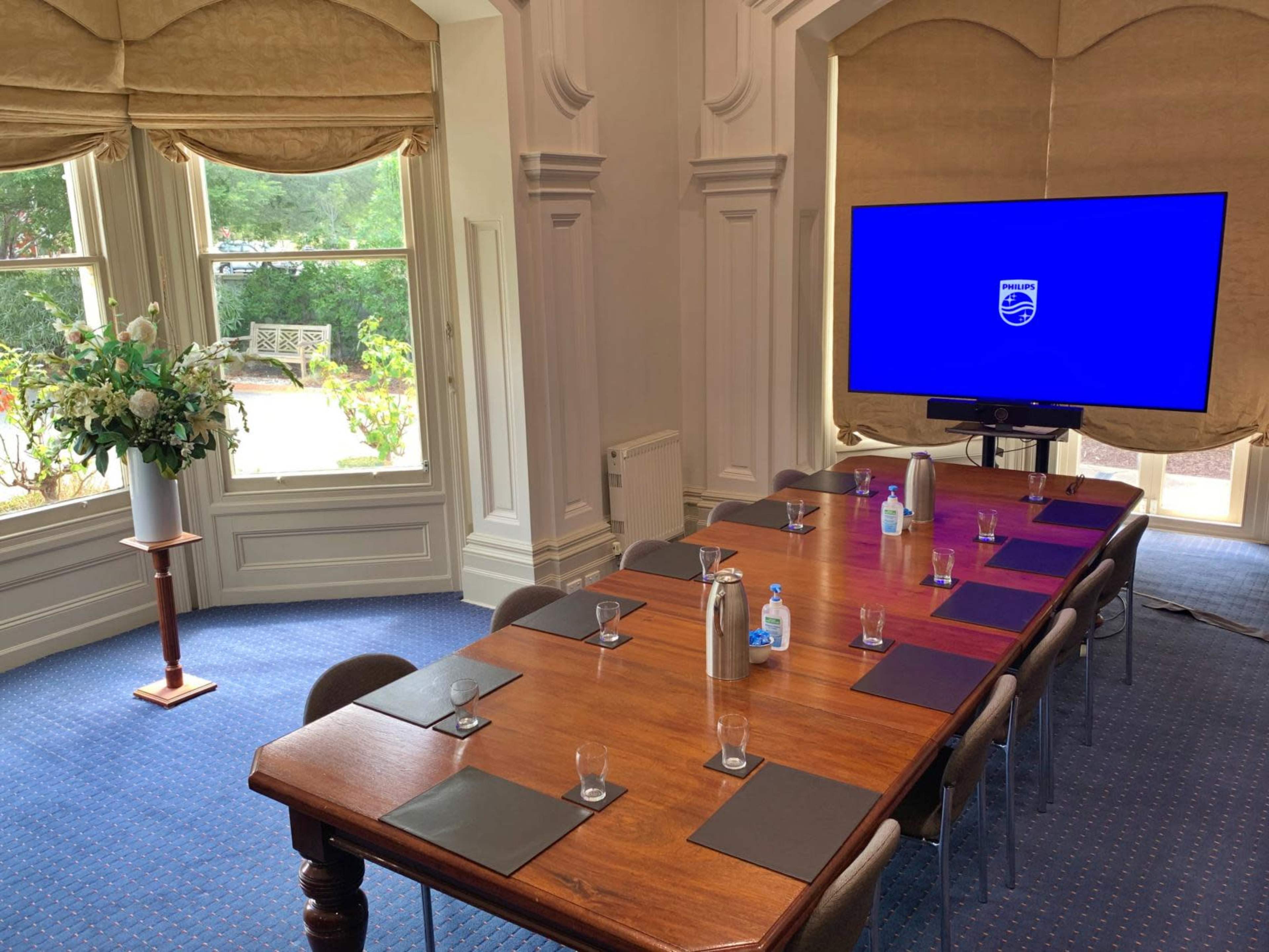 A long wooden conference table is set with water bottles, glasses, and place cards, facing a large screen in a well-lit meeting room with windows overlooking a garden.