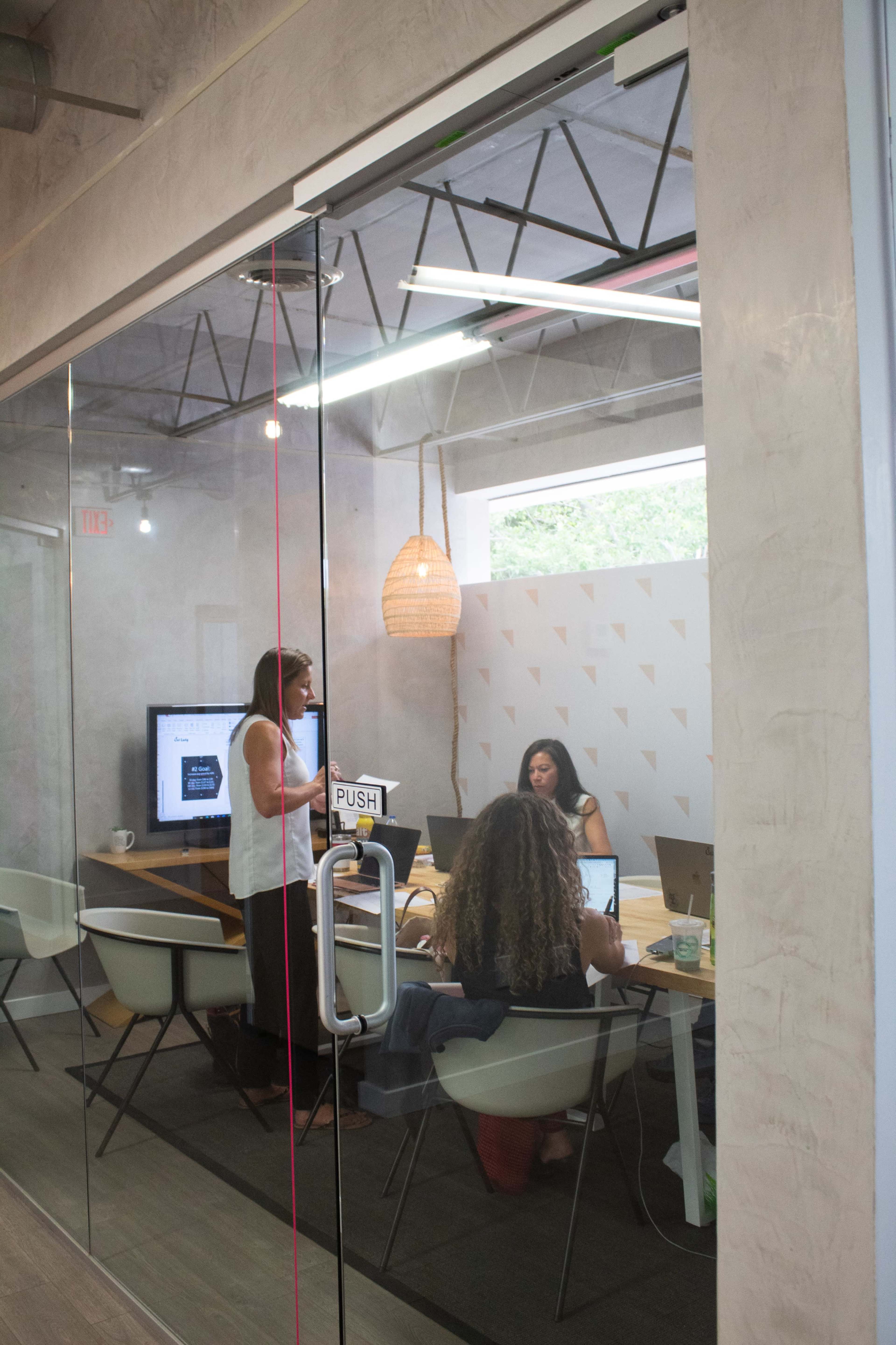 A group of three people is engaged in a meeting in a glass-walled conference room, with laptops and a presentation screen visible.