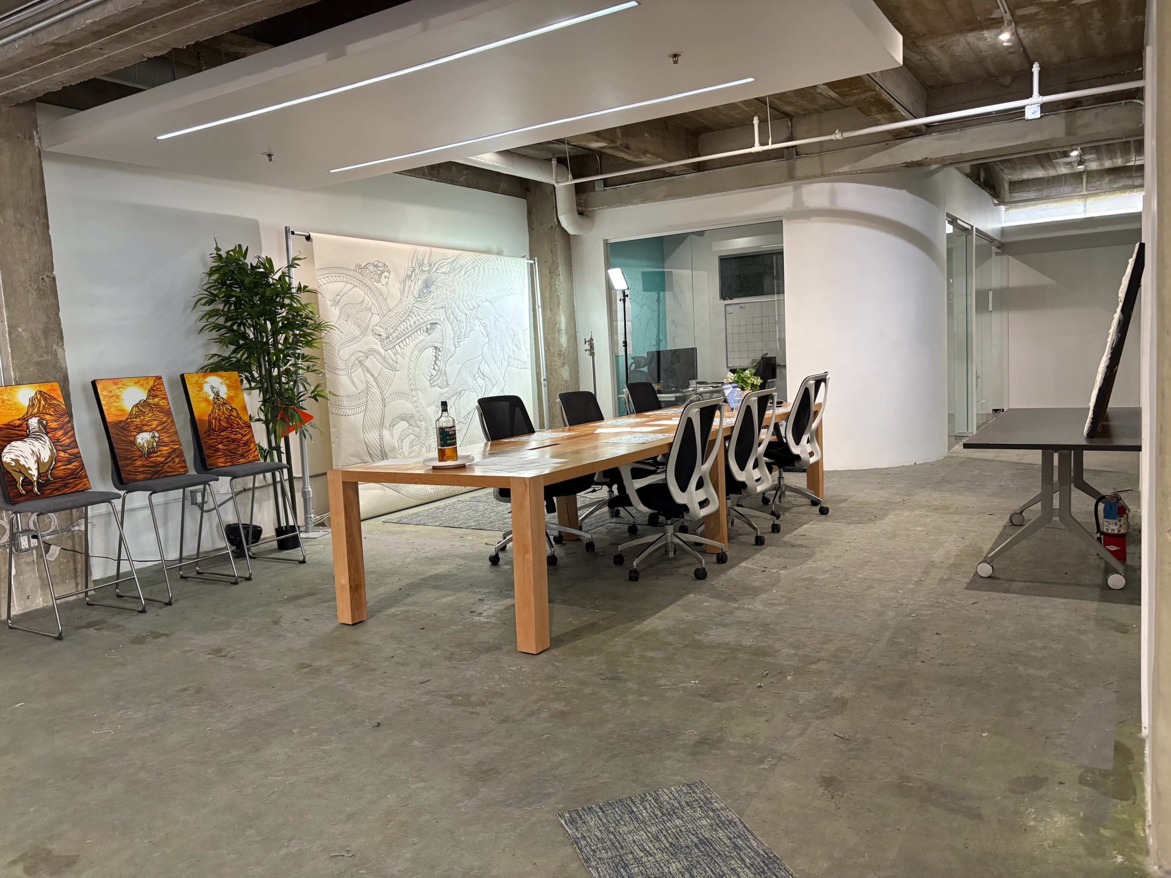 The image shows a spacious office environment featuring a long wooden conference table, several chairs, artwork on display, and an adjacent area with a projector and screens.