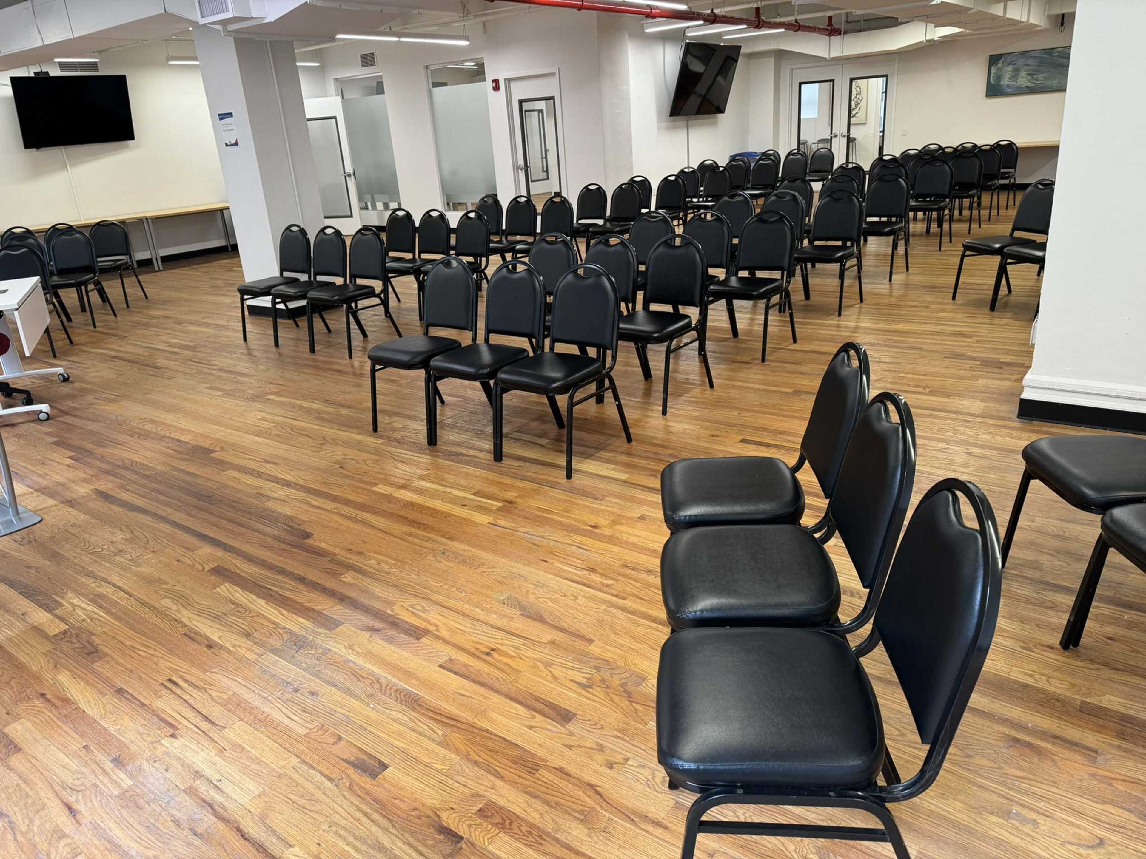The image shows a spacious room with rows of black chairs arranged on a wooden floor.