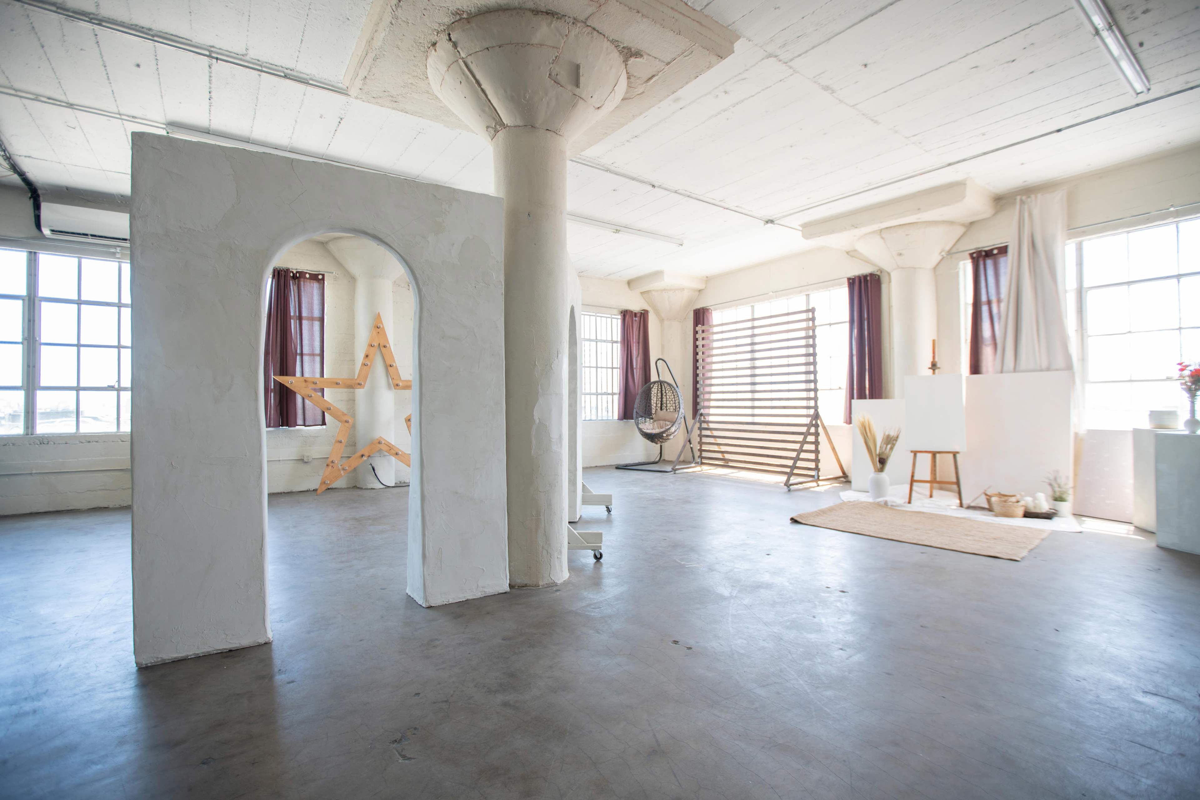 The image shows a spacious, well-lit studio with large windows, featuring minimalist decor, including a wooden star structure and various art supplies.