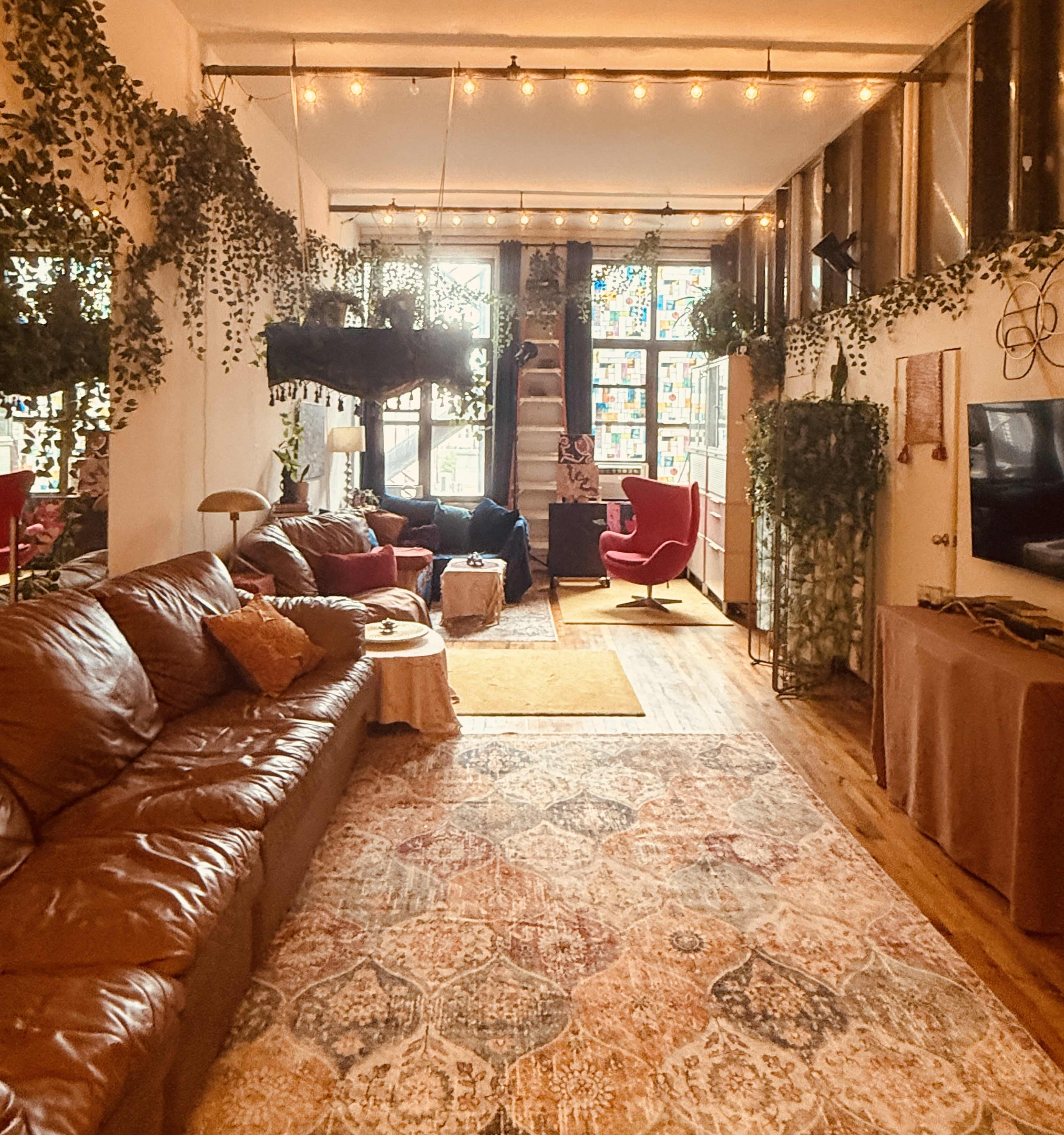 The image depicts a cozy living room with brown leather seating, a colorful area rug, hanging plants, and large windows that allow natural light to flood the space.