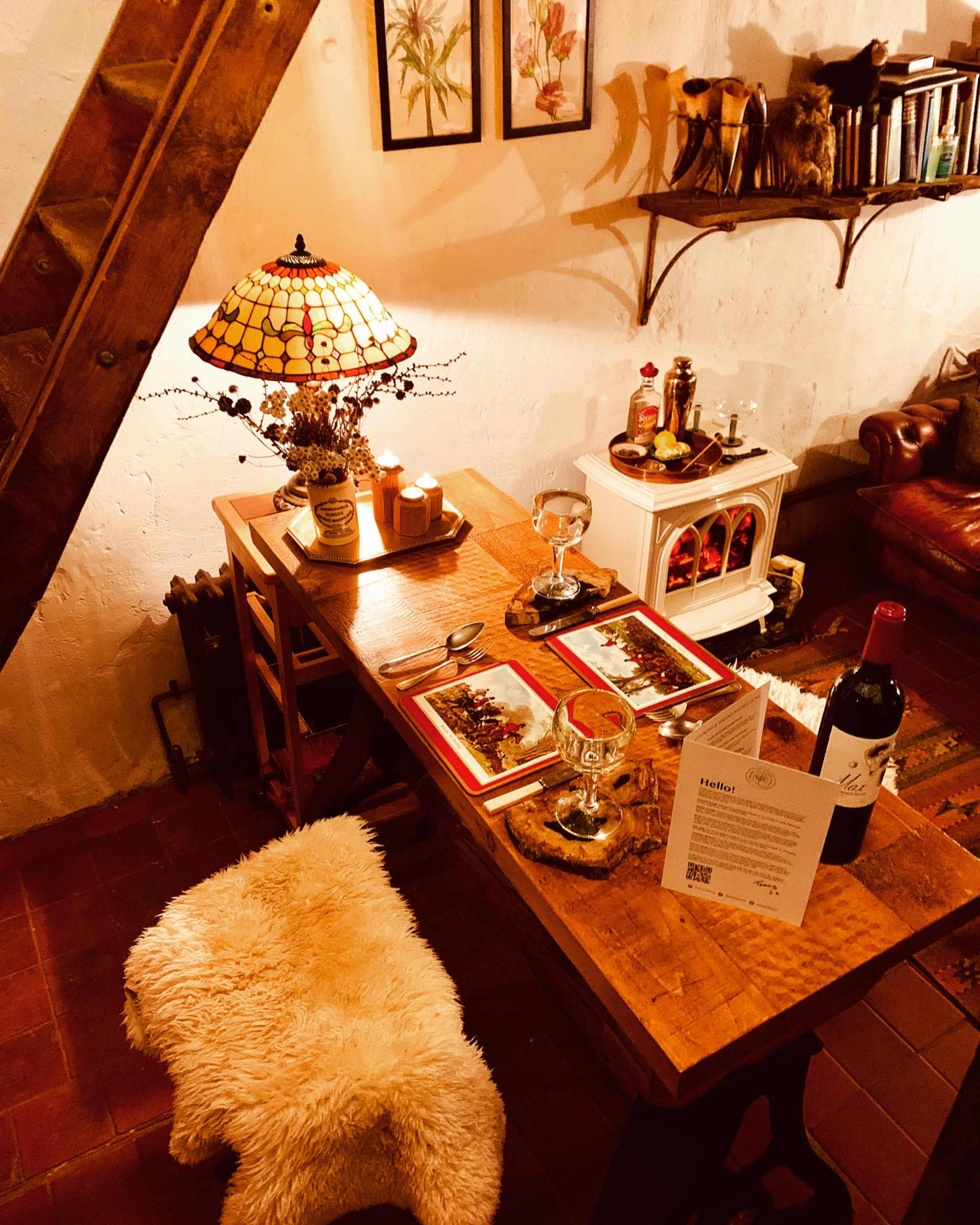 A small wooden table set for two, accompanied by a plush chair, a vintage lamp, and a menu beside a wine bottle in a cozy room.