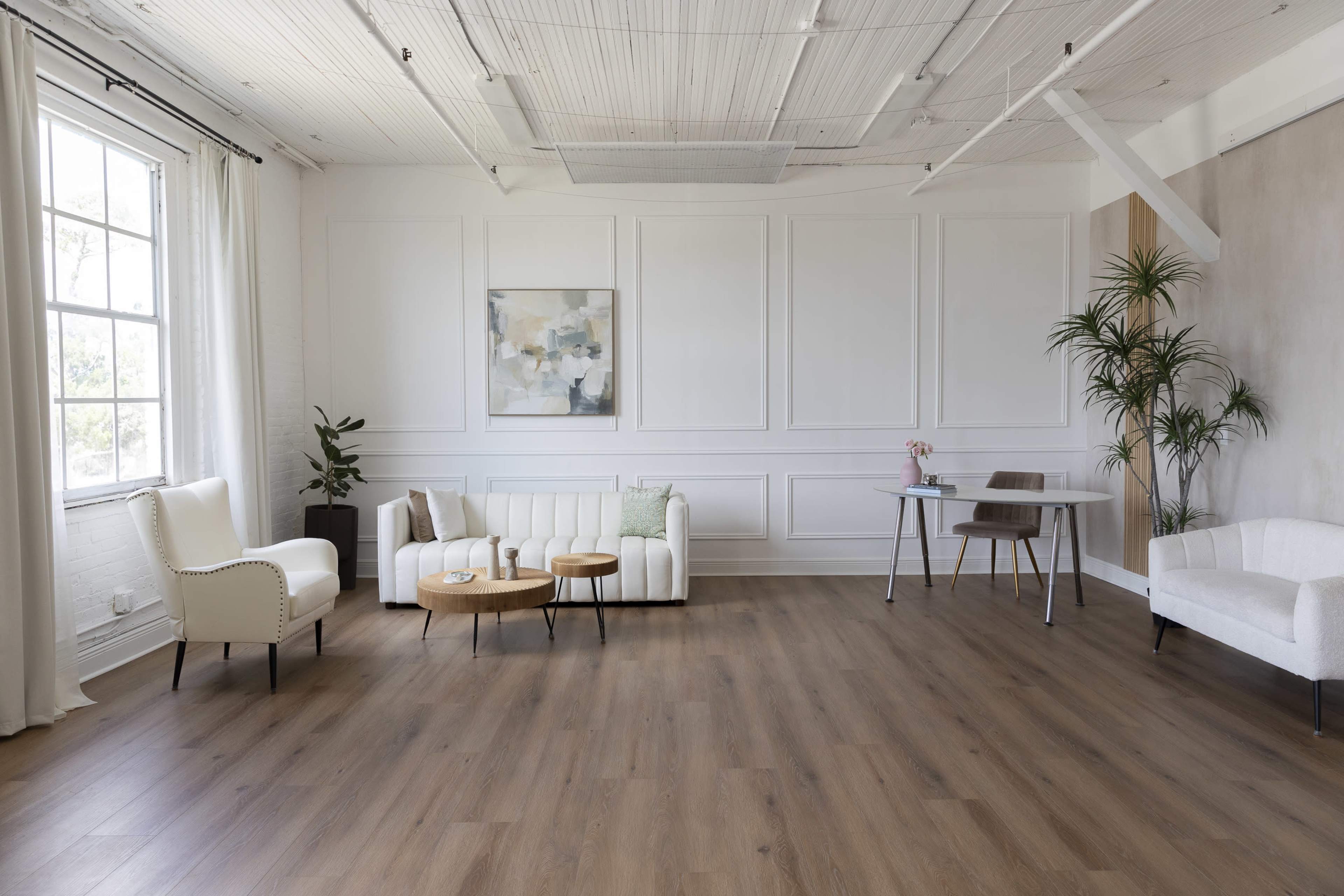 The image shows a bright, modern room with white walls, wooden flooring, and minimal furniture, including a sofa, two chairs, a small table, and a desk with a plant.