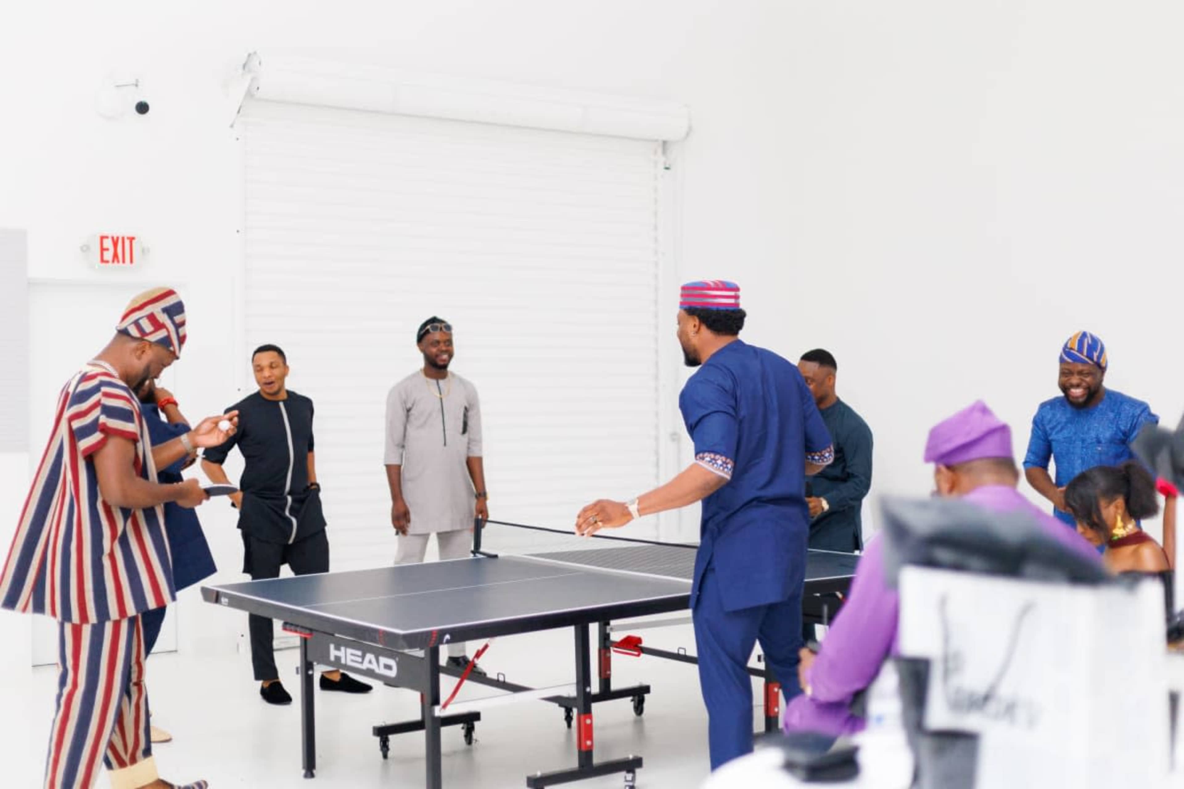 A group of men in various traditional outfits play table tennis in a bright, minimalist indoor space.