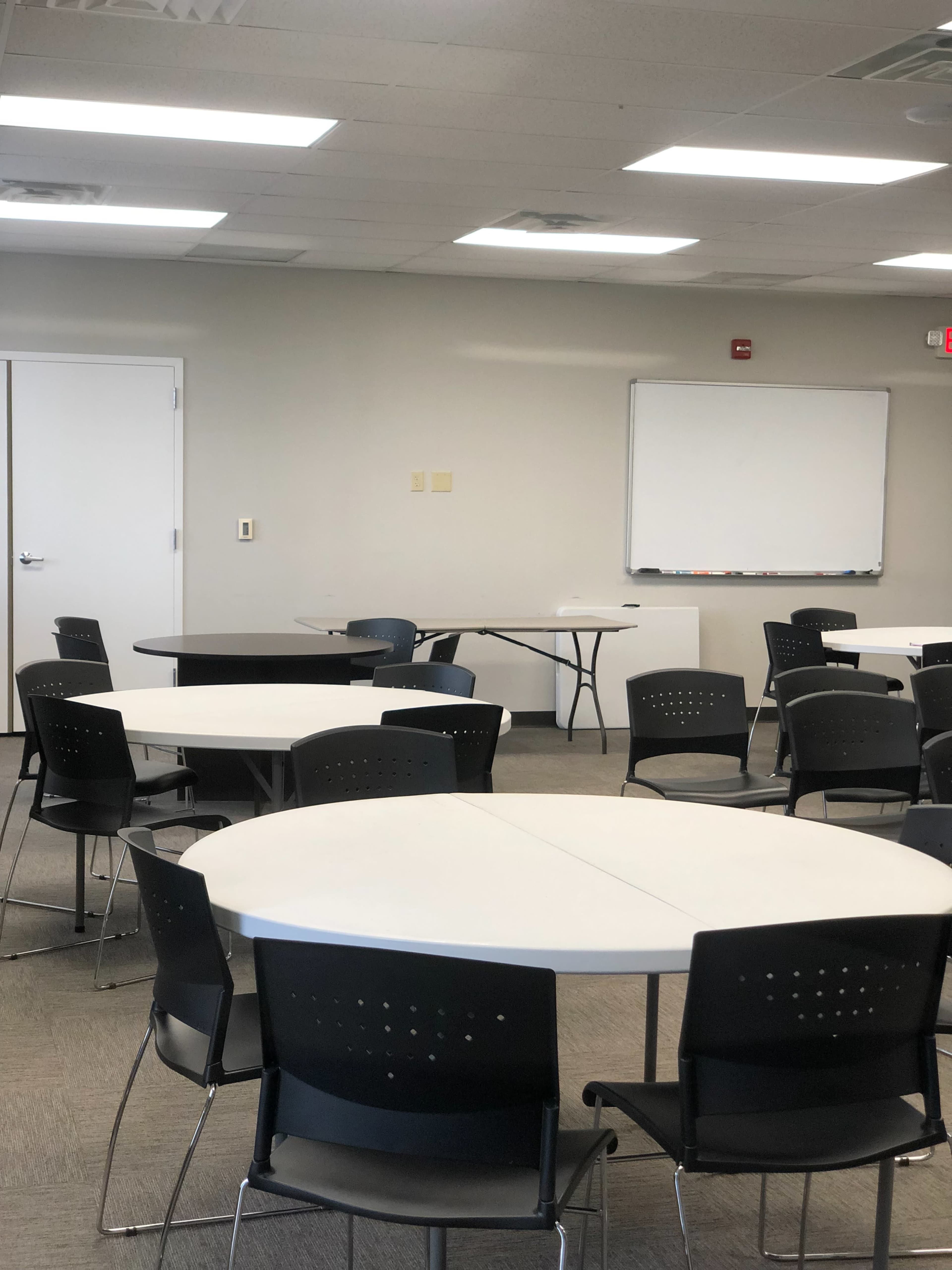 A meeting room with several round tables and black chairs arranged neatly, along with a whiteboard on the wall.